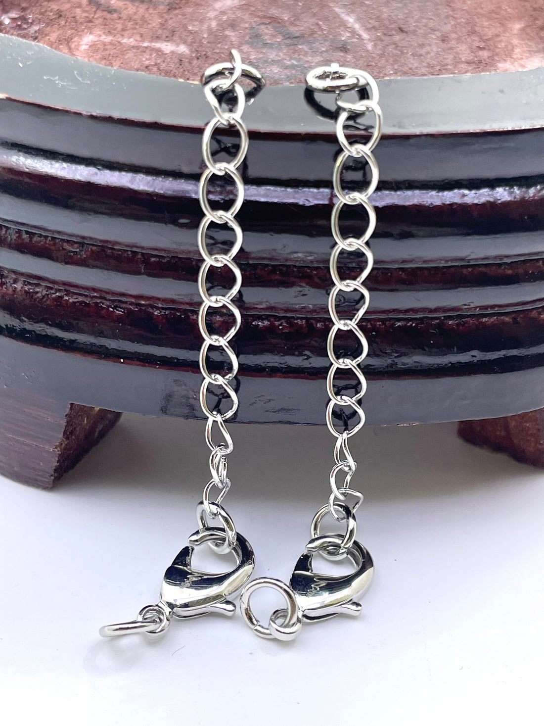 A pair of silver-toned chain extender links with lobster clasp ends, displayed against a dark wooden stand on a white surface.
