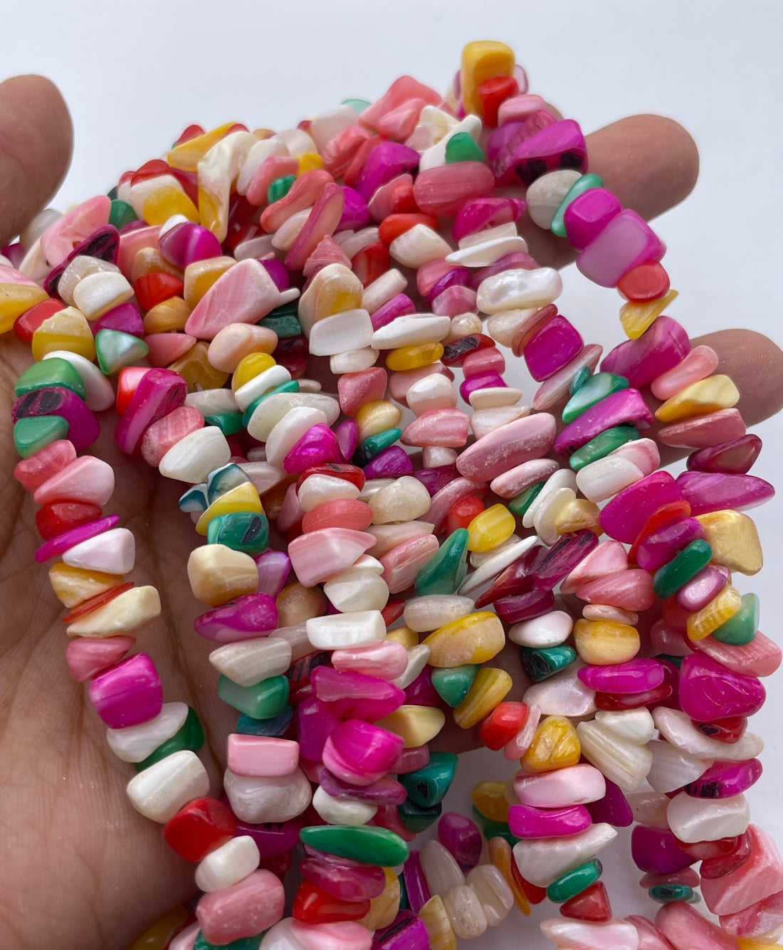 A close-up of a hand holding a pile of colorful, irregularly shaped polished mop stone beads on strands, set against a plain white background.