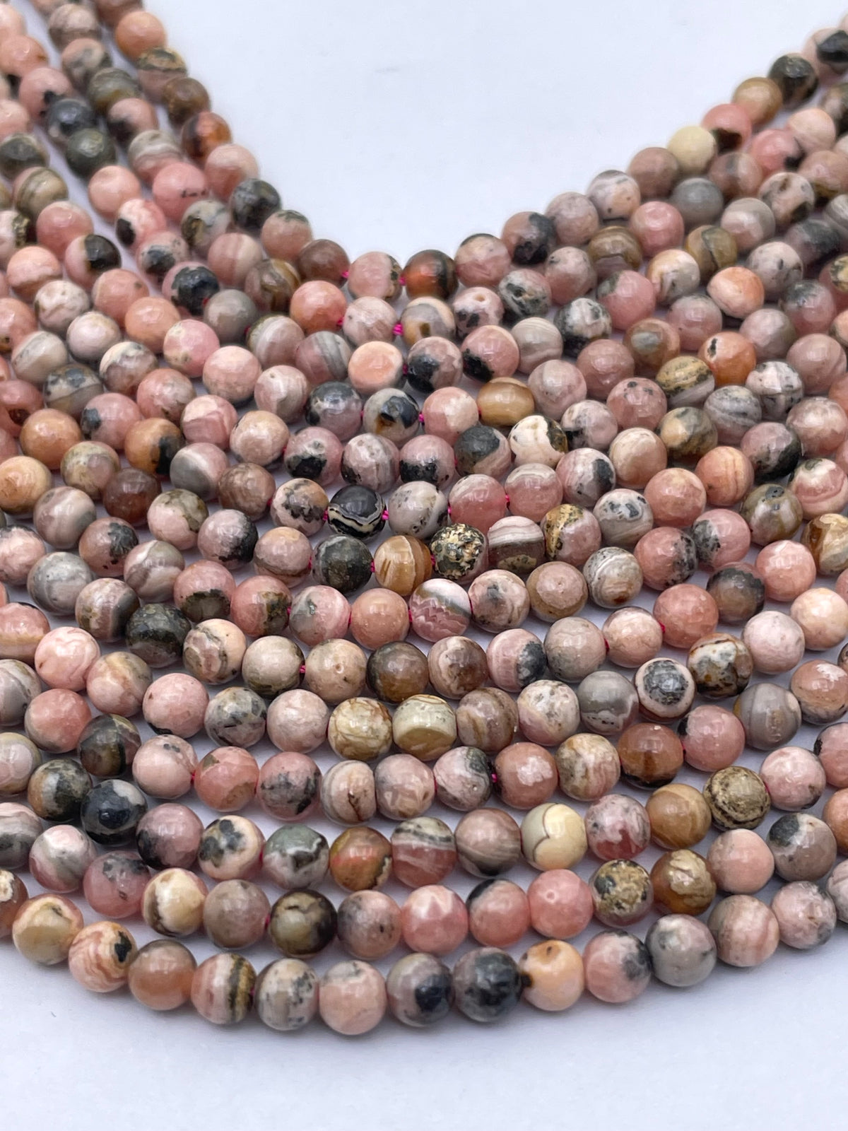 Multiple strands of round polished rhodochrosite beads in pink, peach, gray, black, and beige marbled stone, arranged in overlapping curves on a white background.