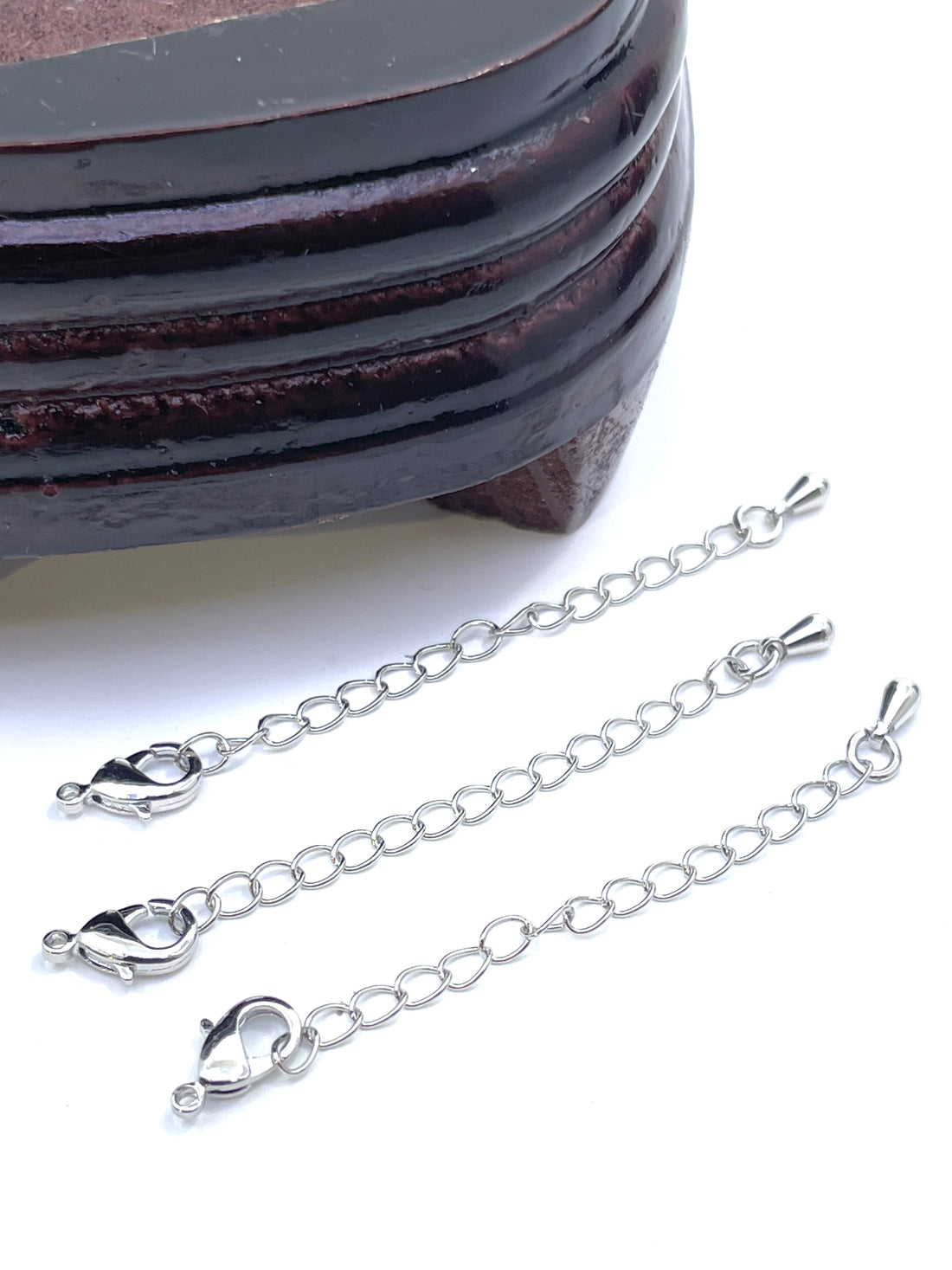 Three silver-tone chain extender links with lobster clasp ends, arranged diagonally on a white background next to a dark wooden block.