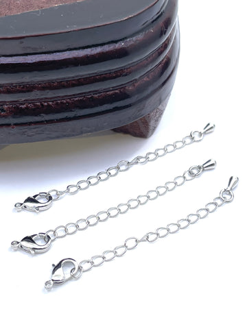 Three silver-tone chain extender links with lobster clasp ends, arranged diagonally on a white background next to a dark wooden block.