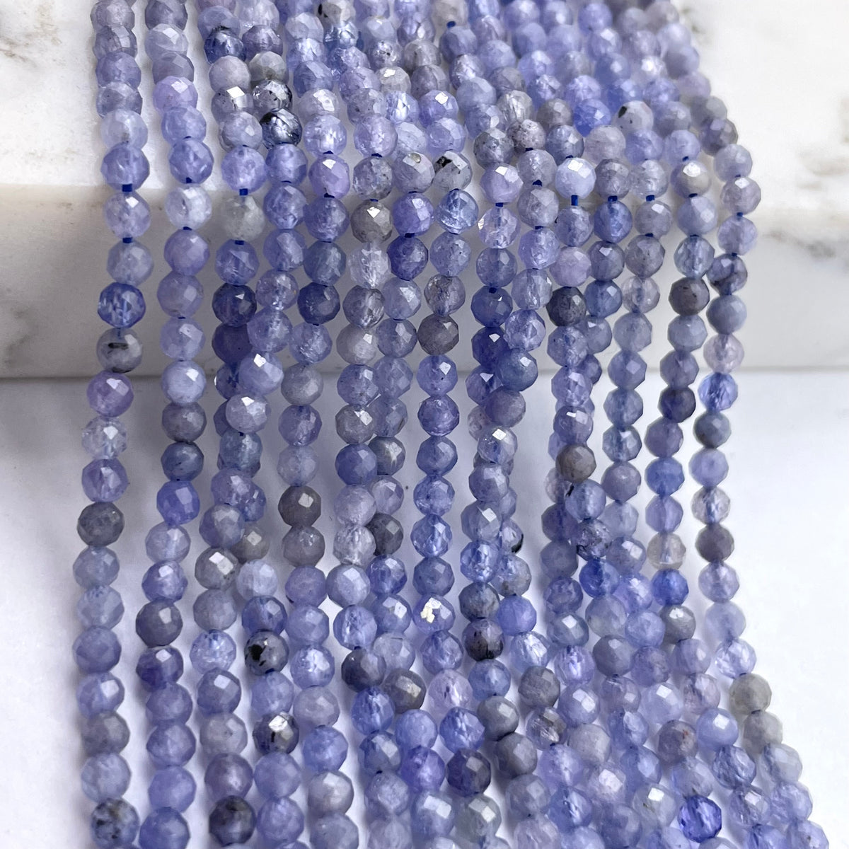 Multiple strands of faceted round tanzanite beads in shades of periwinkle, lavender, and slate blue, draped over a white marble surface.