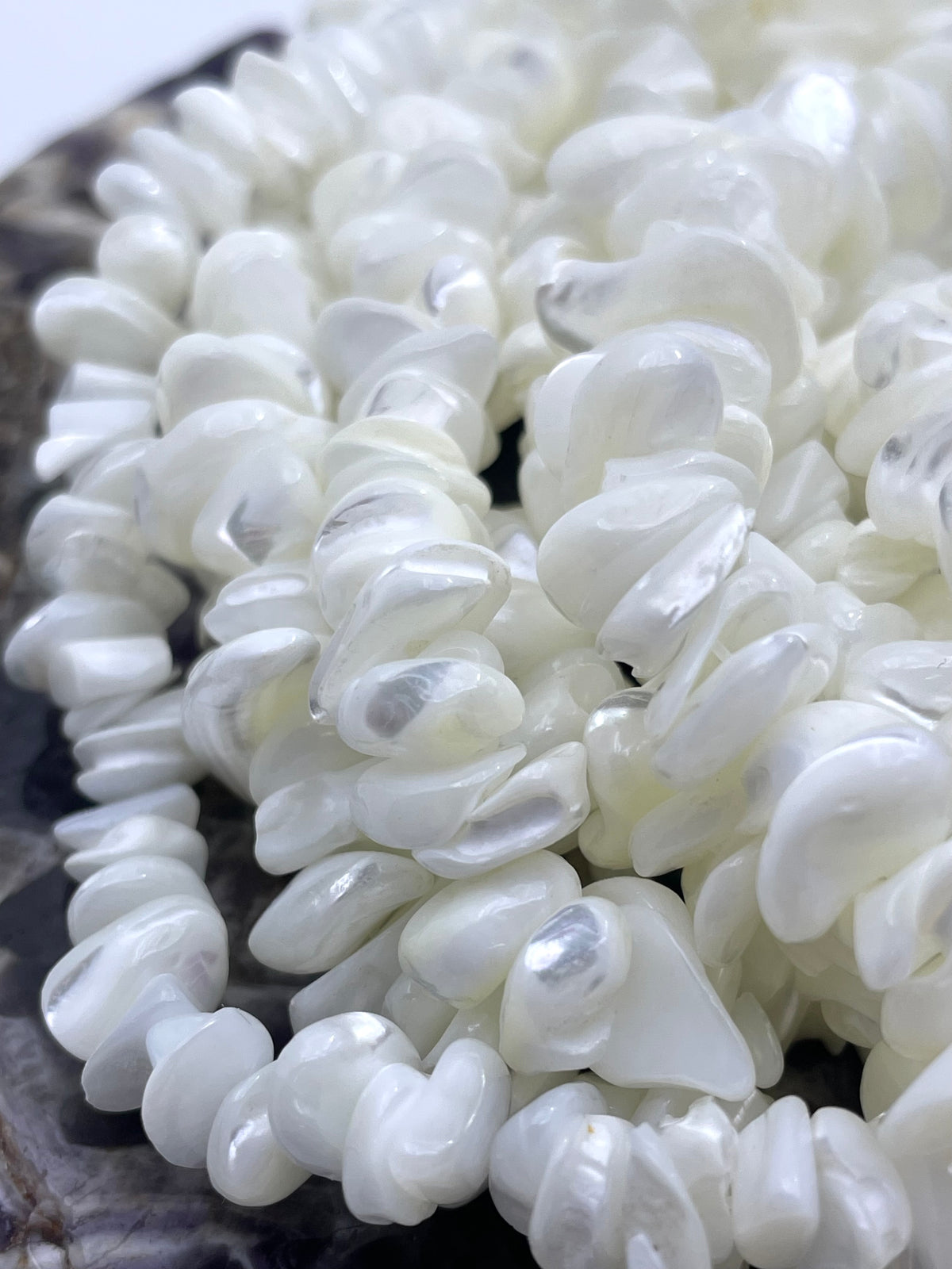 A close-up of a strand of white freshwater pearl chips with a glossy, lustrous finish, resting on a dark textured surface.
