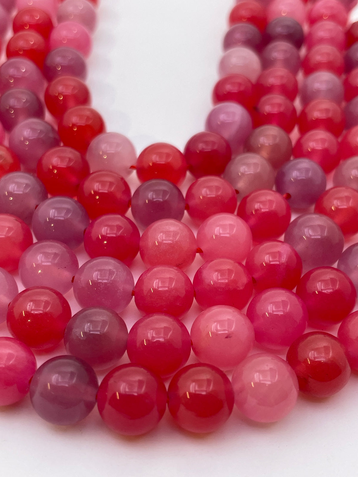 Close-up of multiple strands of round polished scarlet agate beads in shades of pink, red, and purple on a white background.