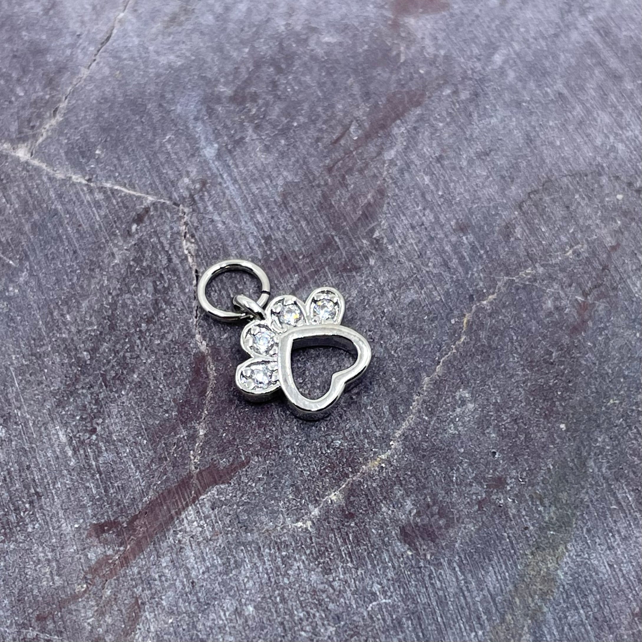 A silver-colored metal charm shaped like a paw print with a heart outline, adorned with small clear rhinestones, resting on a textured gray slate surface.