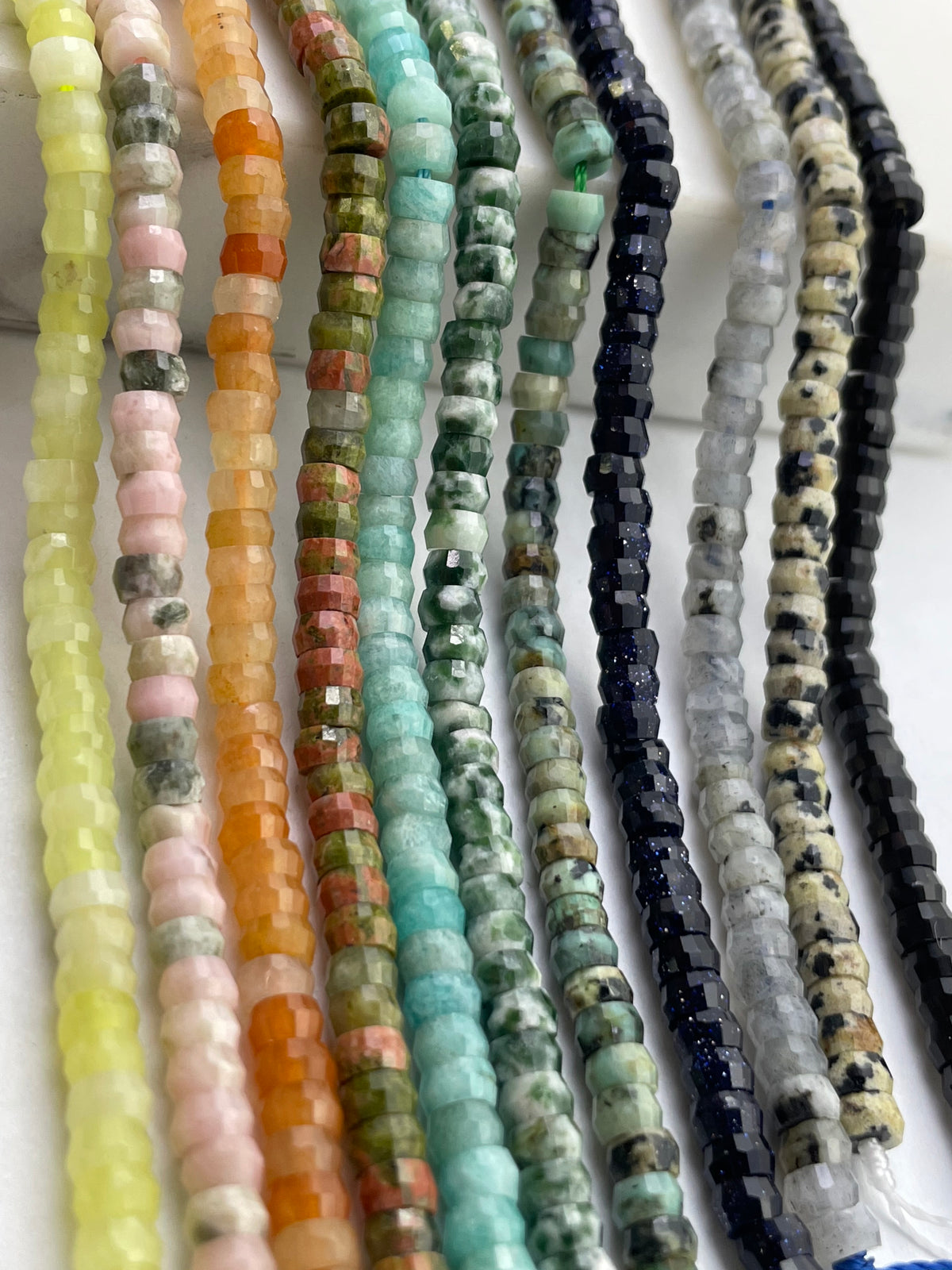 Multiple strands of faceted gemstone beads in assorted colors and patterns, arranged vertically on a light background.