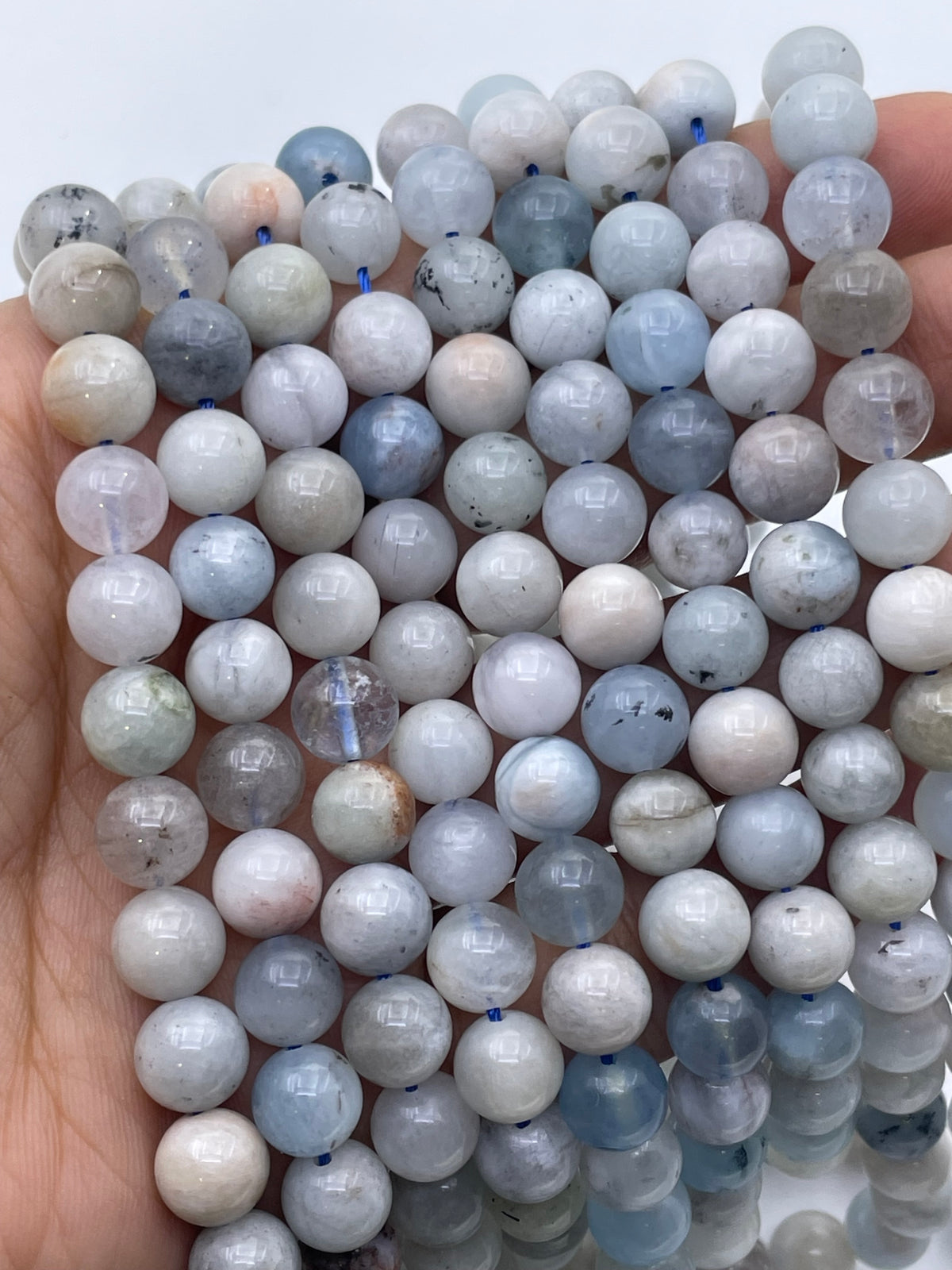 A close-up of a hand holding a bundle of polished round aquamarine gemstone beads in pale blue, gray, white, and soft pink tones against a plain light background.