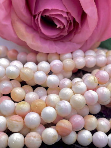 Close-up of strands of round queen conch shell pearl beads in soft white, blush pink, peach, and cream tones, with a large pink rose in the background.