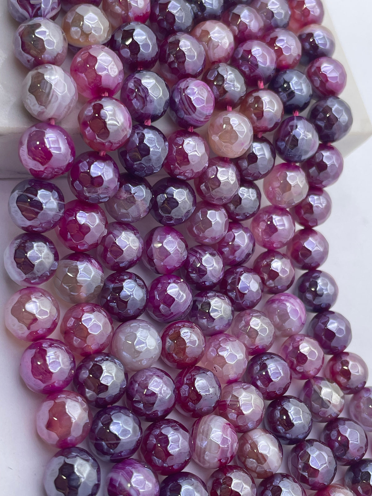 Faceted round pink agate gemstone beads in shades of pink, purple, and white, arranged in diagonal rows on a light background.