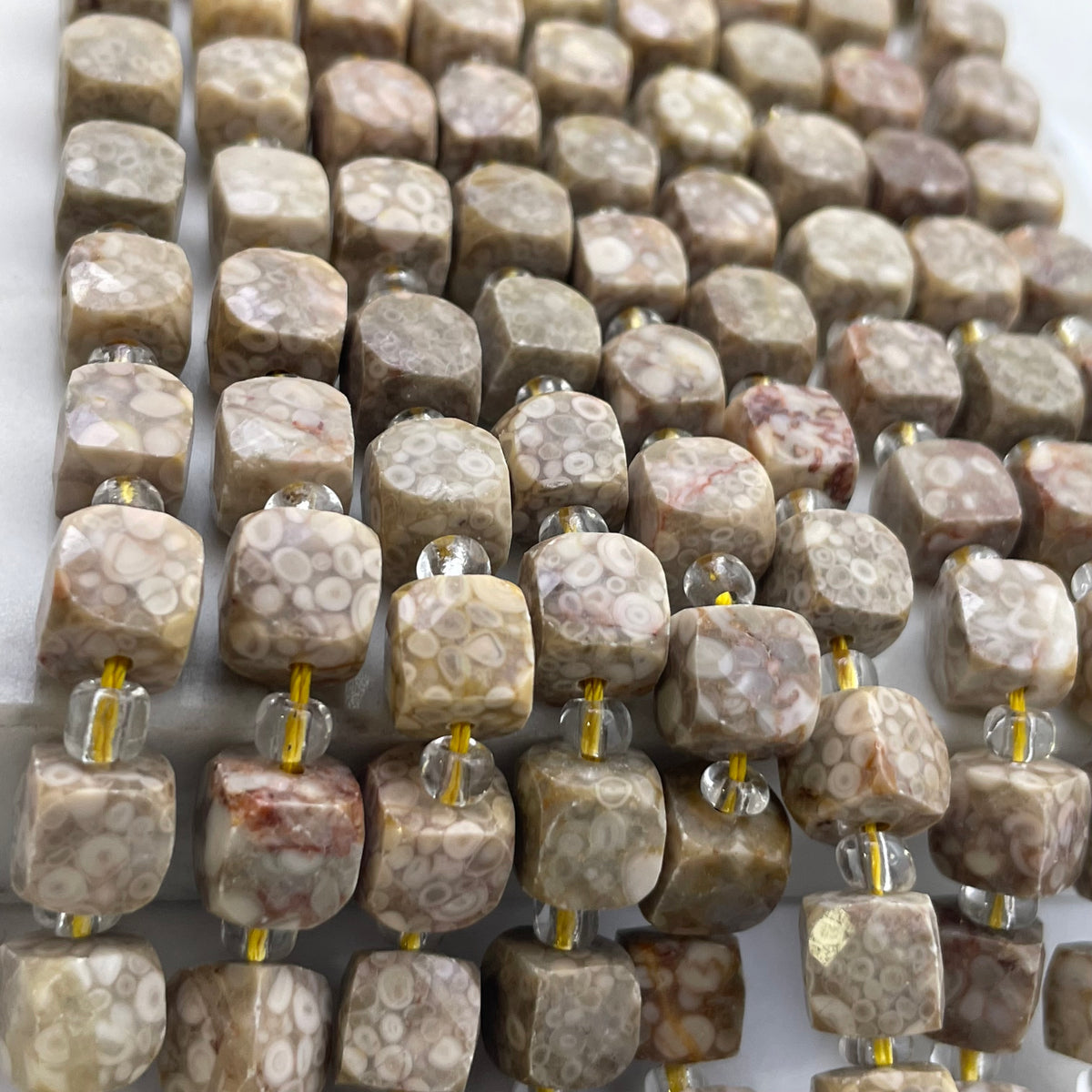 A close-up of multiple strands of faceted square beads made from a beige and white marbled stone with organic circular patterns, threaded on clear glass beads with small yellow spacer accents.