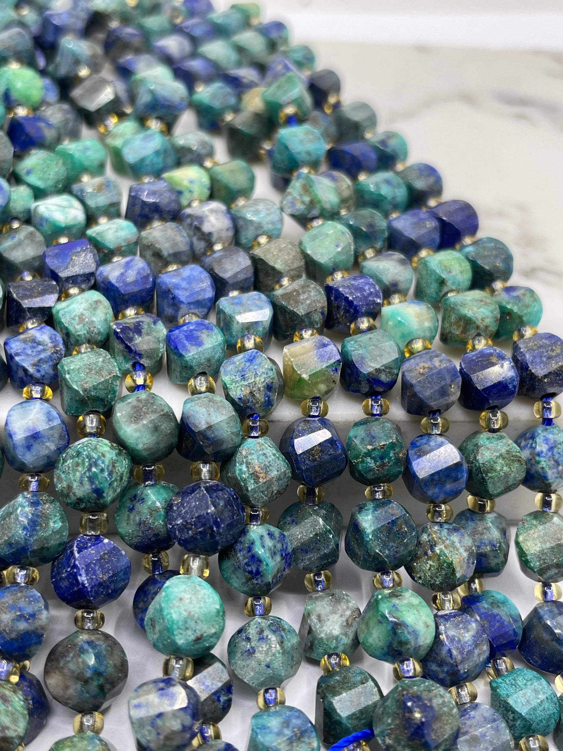 Close-up of faceted chrysocolla lapis gemstone beads strung in multiple strands, showing blue, green, teal, and gray stones separated by small metallic spacer beads.