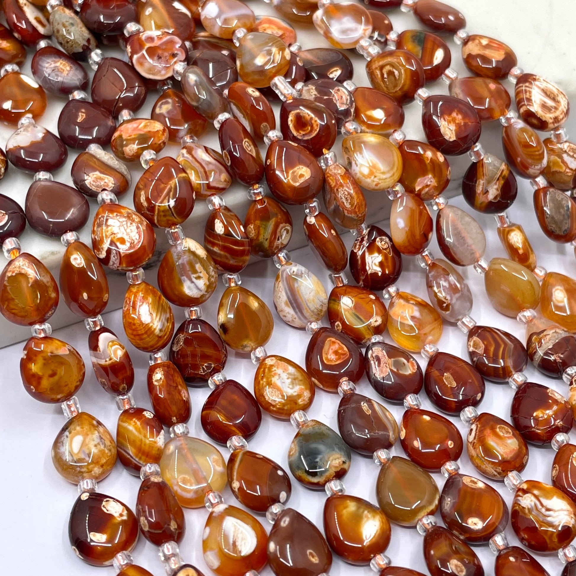 A close-up of a strand of polished teardrop-shaped terracotta beads in various earthy brown, amber, and reddish-orange patterns, separated by small silver spacer beads.