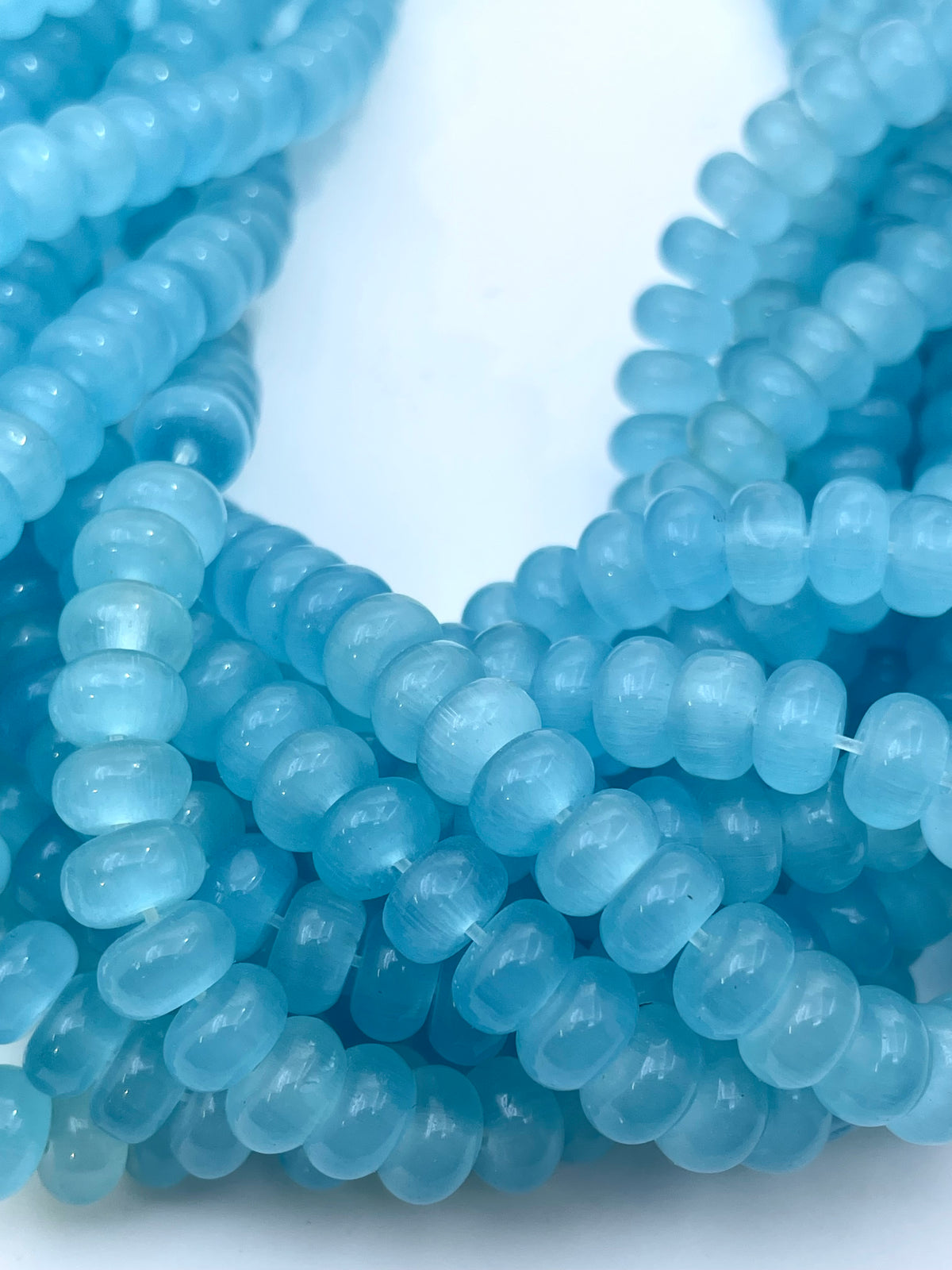 Multiple strands of light blue cats eye translucent smooth donut beads arranged on a white background.
