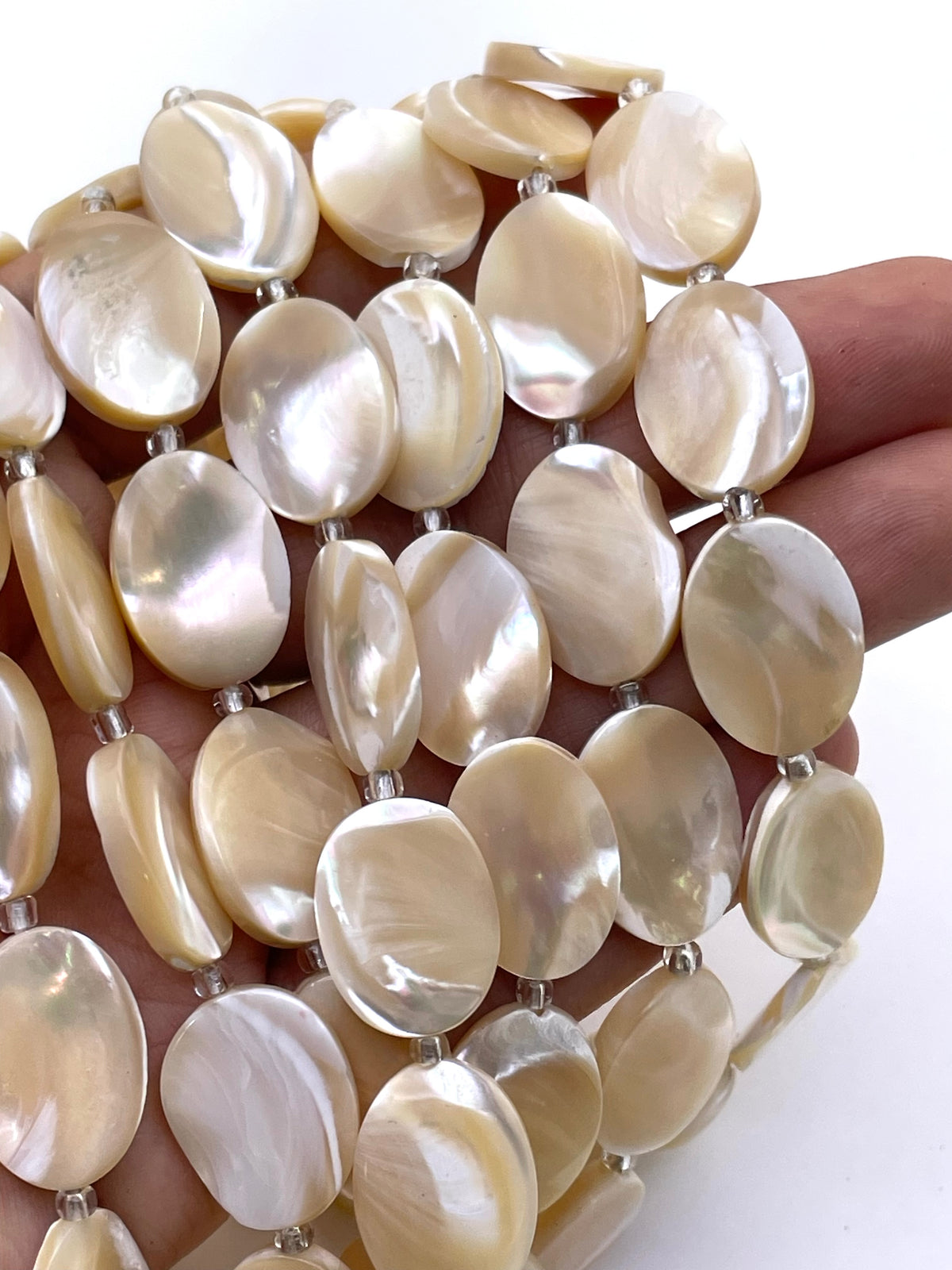 A close-up of a hand holding a strand of oval mother-of-pearl beads with a glossy, iridescent finish, set against a plain white background.