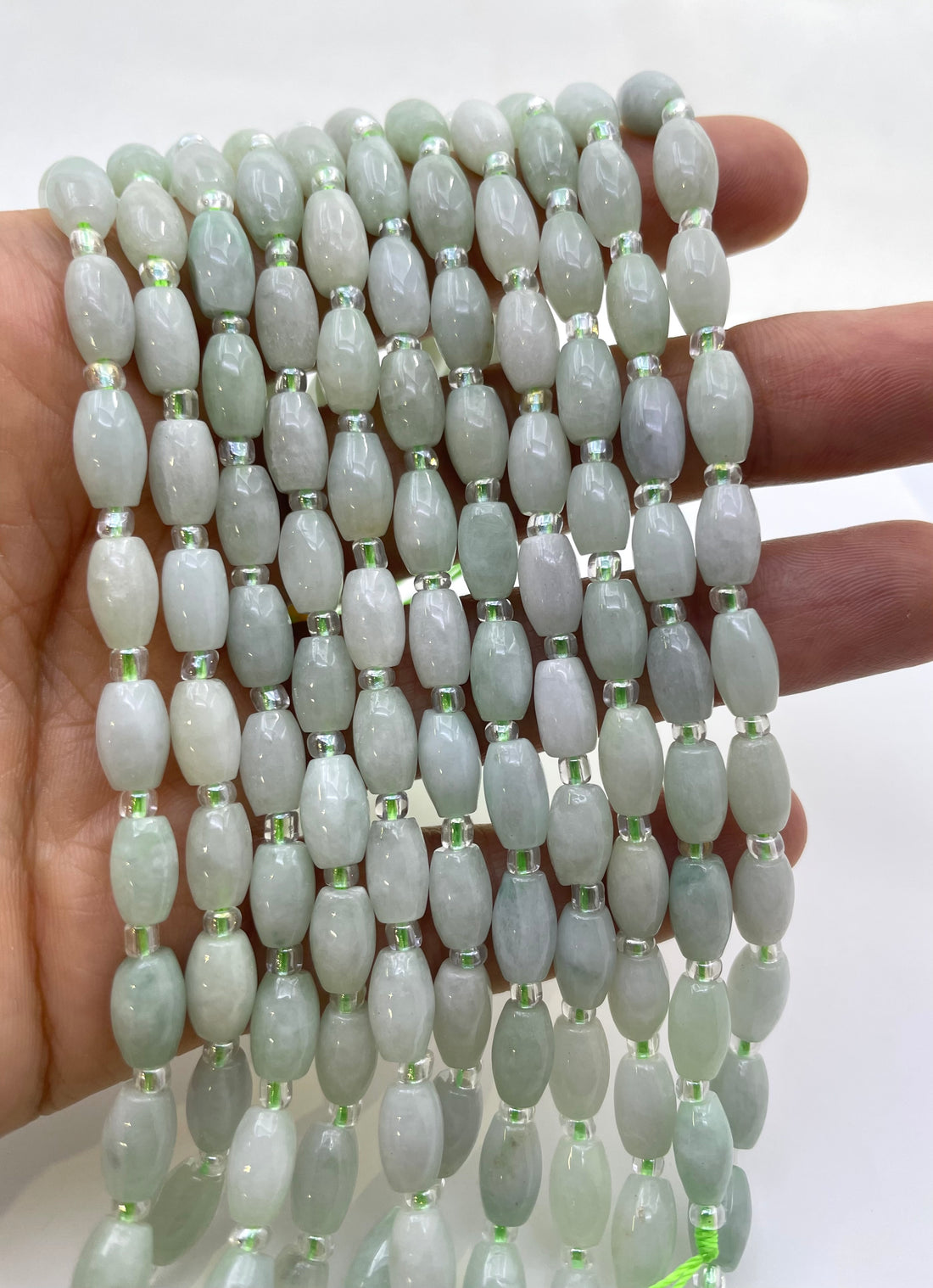 A hand holding multiple strands of  light green burma jade beads with small metallic spacer rings, shown against a plain white background.