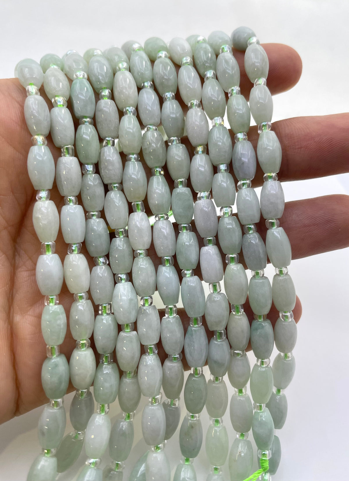 A hand holding multiple strands of  light green burma jade beads with small metallic spacer rings, shown against a plain white background.