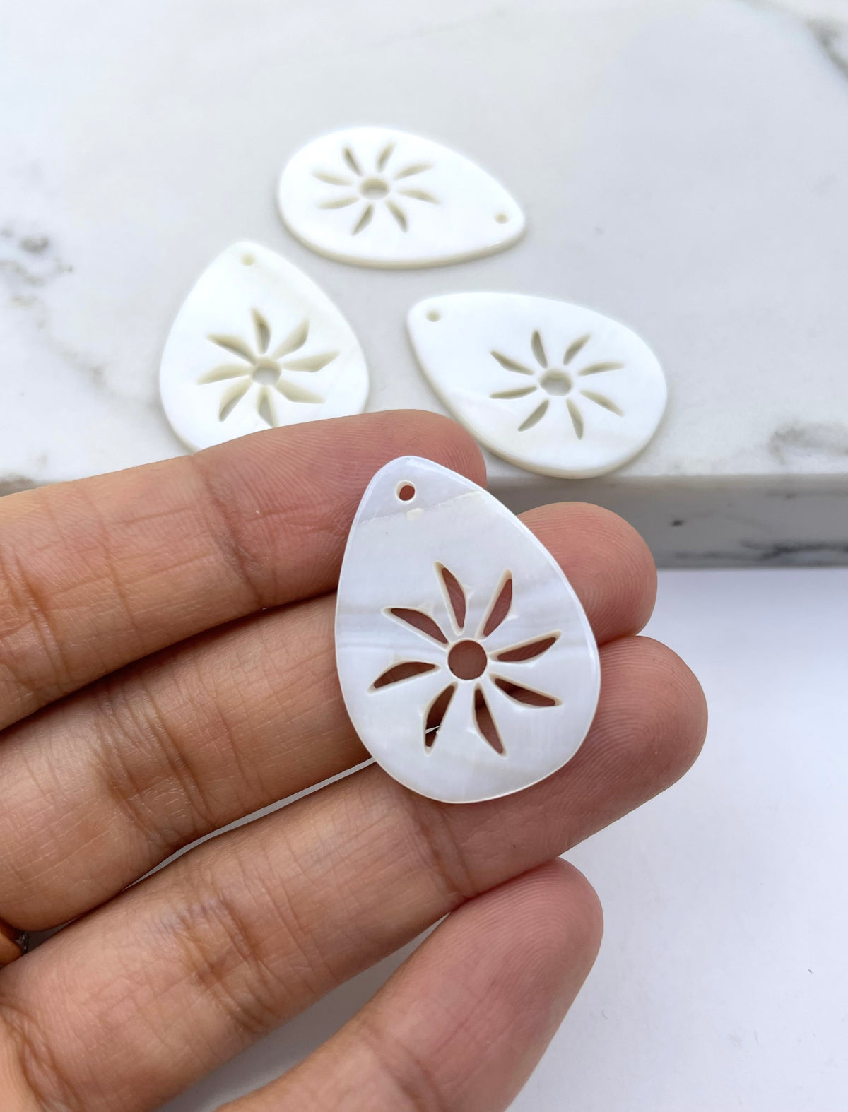 A hand holds a small white teardrop-shaped shell charm with a sand dollar cutout pattern, with three similar charms blurred in the background on a light marble surface.