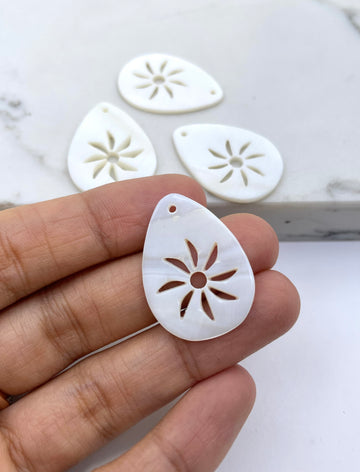 A hand holds a small white teardrop-shaped shell charm with a sand dollar cutout pattern, with three similar charms blurred in the background on a light marble surface.