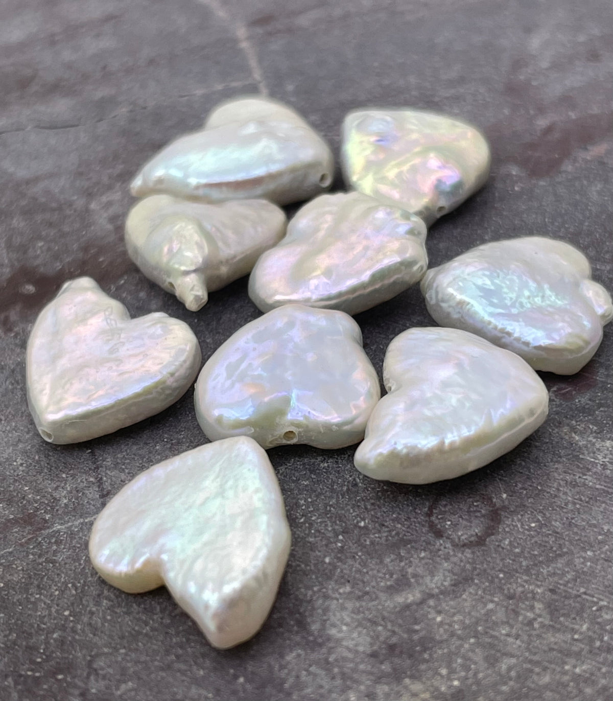 Nine irregular heart-shaped baroque pearls with iridescent white and pale green hues, arranged on a dark textured stone surface.