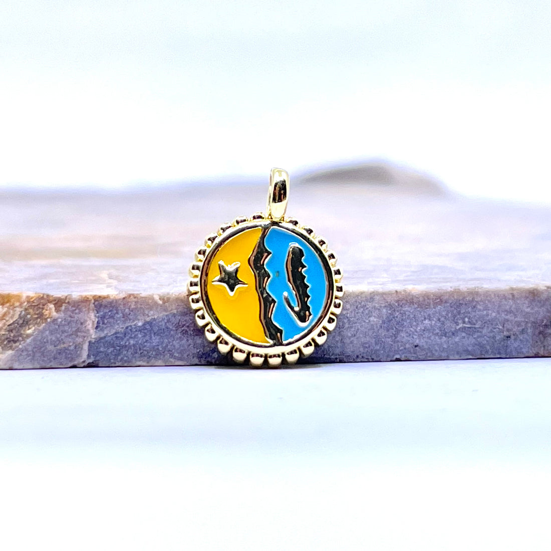 A round gold-toned pendant charm featuring a split yellow and blue design with a black silhouette and star, resting on a gray stone surface against a white background.