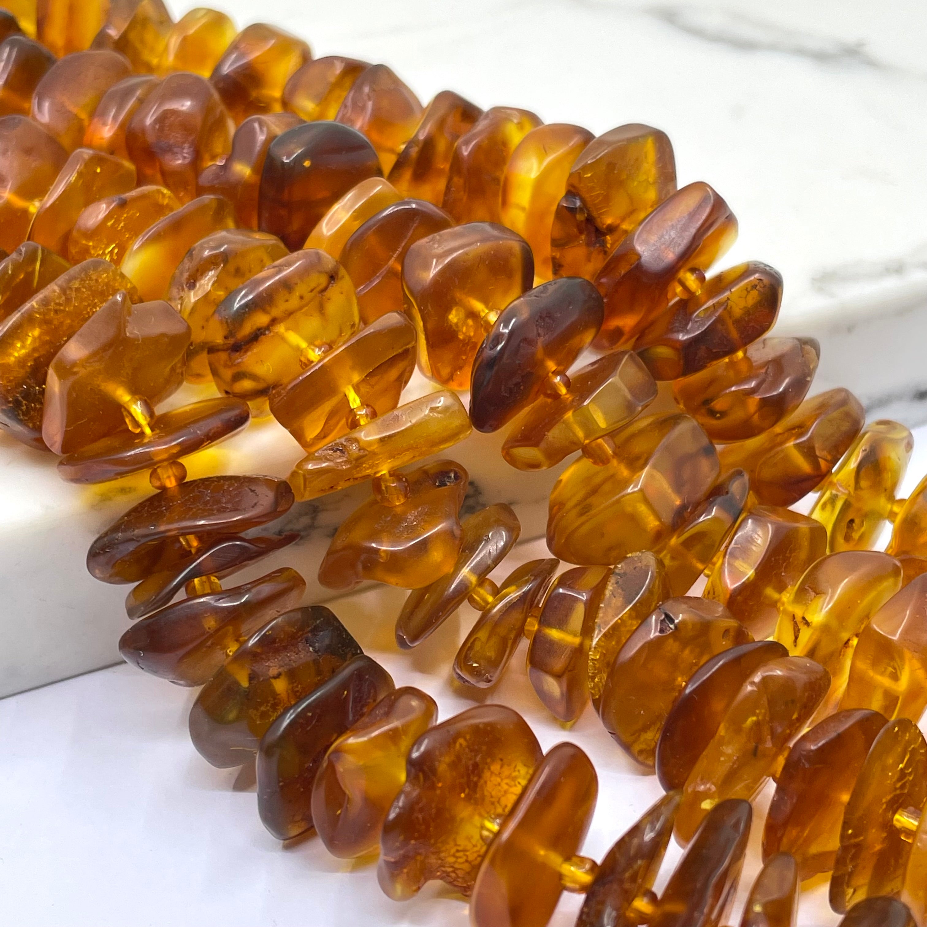 Close-up of amber chip beads strung on a cord, showing translucent warm brown and honey-gold tones with natural inclusions, photographed on a light marble-like surface.