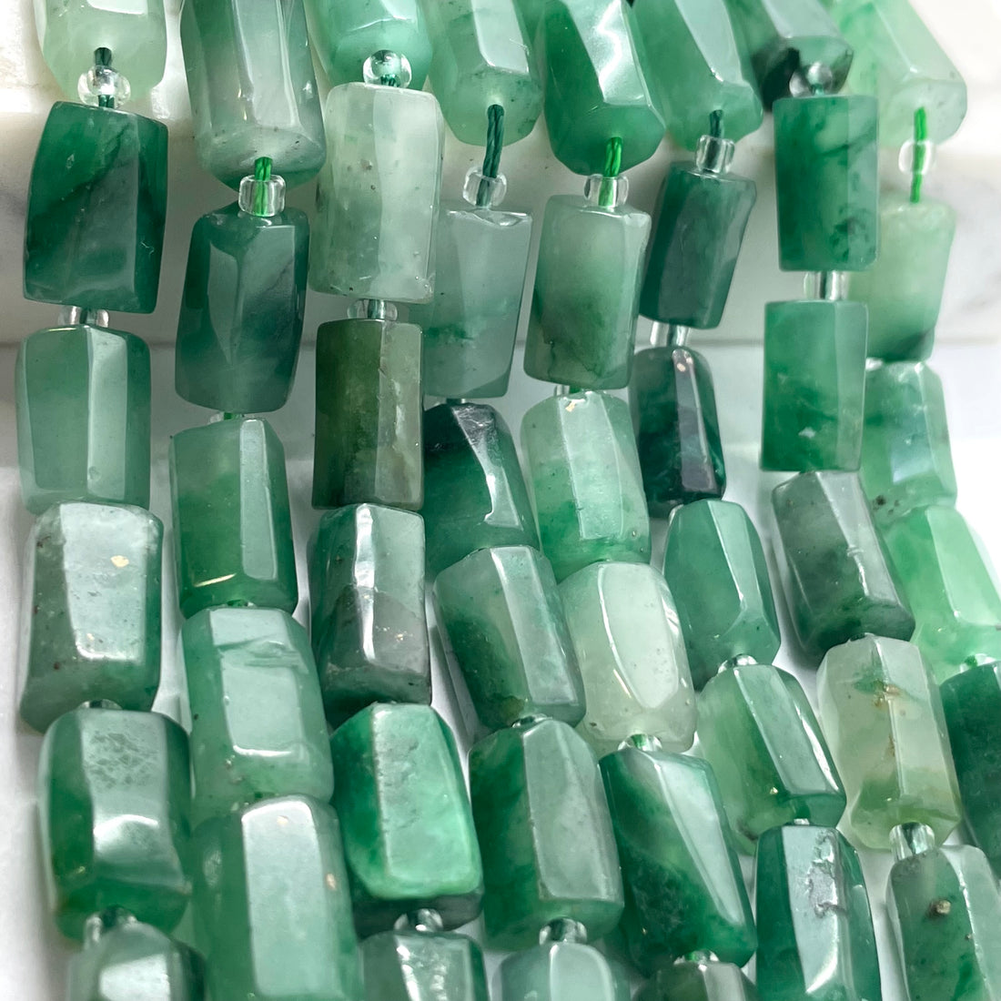 A close-up of a hand holding a strand of rectangular green jade stone beads with a polished, faceted finish.