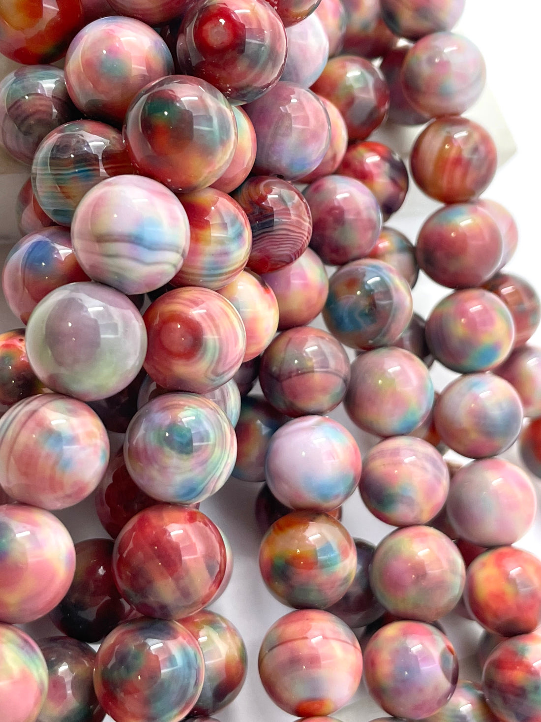 A close-up of glossy round fall foliage agate beads with marbled swirls of pink, red, blue, purple, and white on a white background.