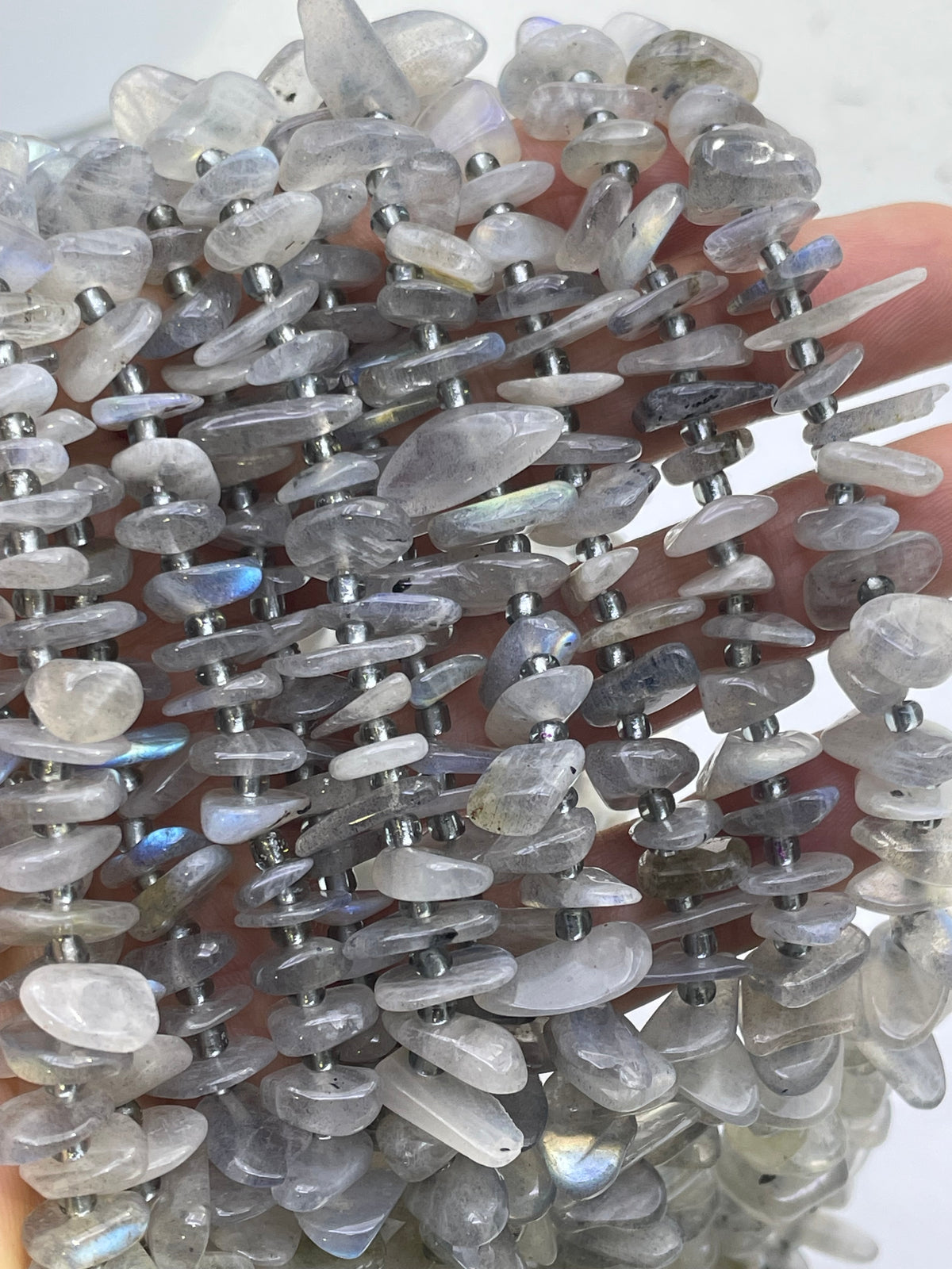 A close-up of a strand of gray labradorite tumbled stone chips with silver spacer beads, held in a hand against a light background.