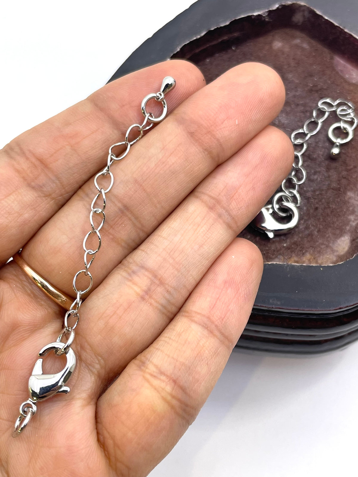 A close-up of a hand holding a silver chain extender bracelet with a lobster clasp, with a dark jewelry box in the background.