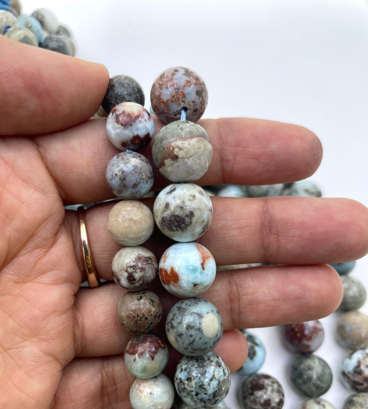 A close-up of a hand holding a strand of round natural stone larimar beads. The beads have a mottled pattern of light blue, gray, white, brown, and black, with one bead showing a light blue center and orange-brown rim.