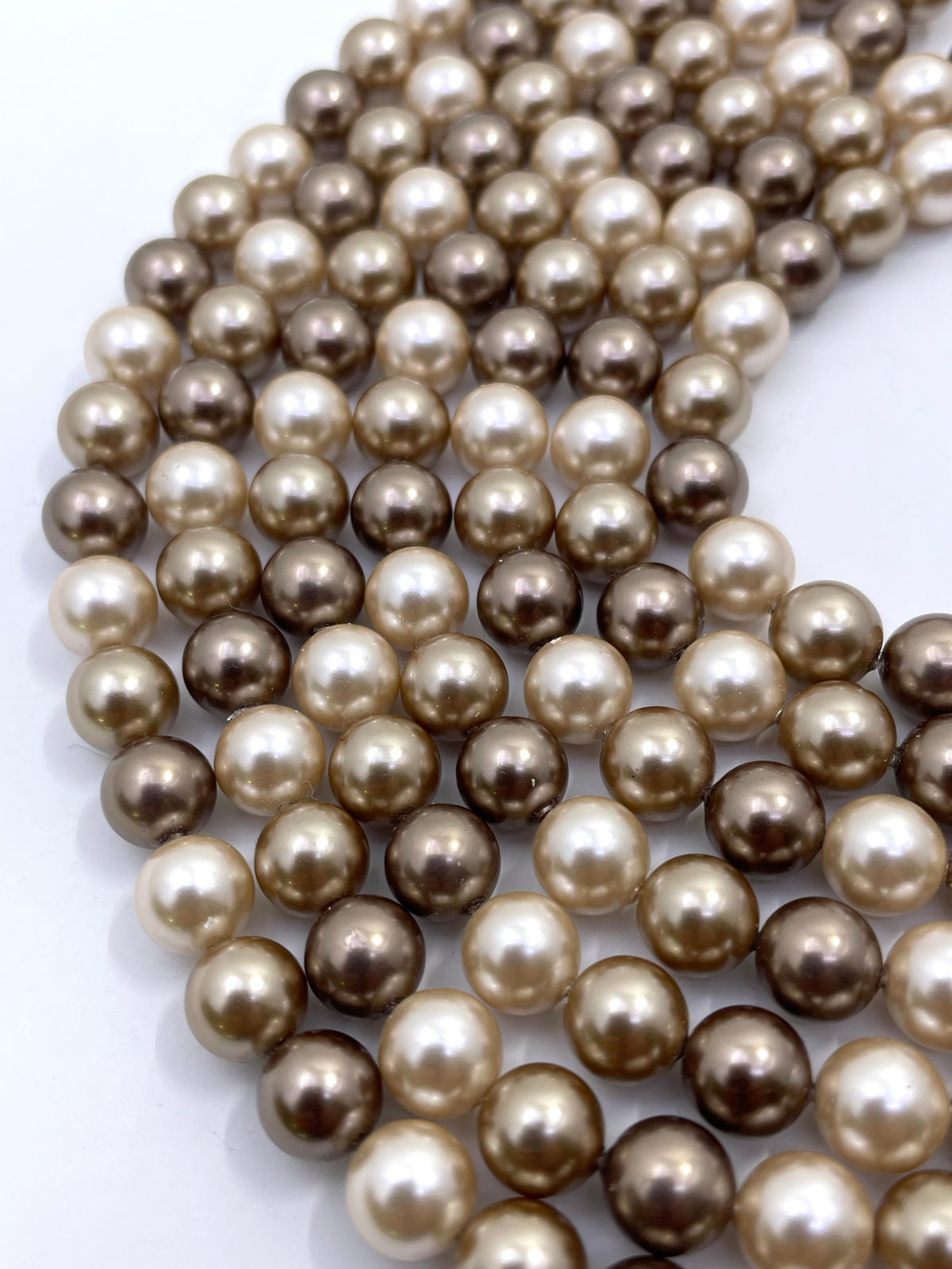Multiple strands of round shell pearl beads in a mix of champagne, beige, and dark brown tones, arranged on a plain white background.