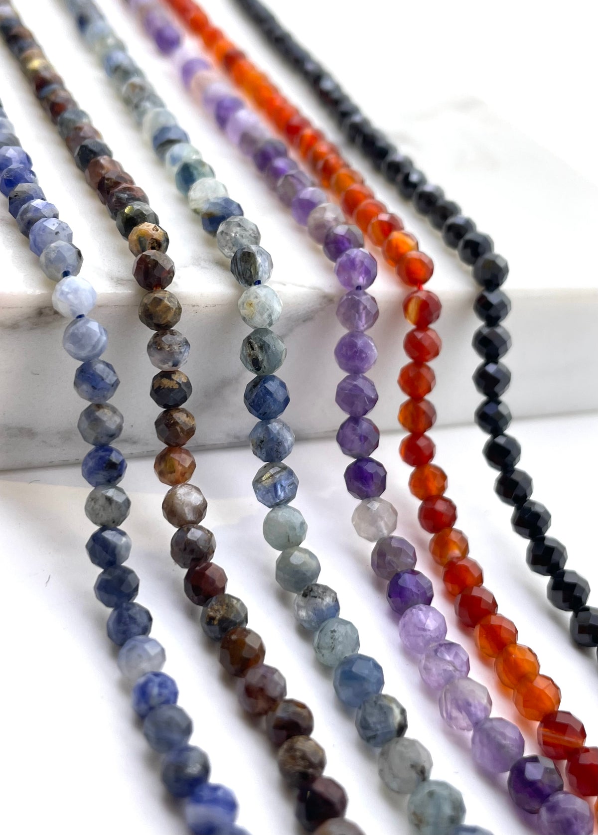 Multiple strands of faceted round gemstone beads in assorted colors, including blue, gray, brown, orange, purple, and black, arranged diagonally on a white marble block against a white background.