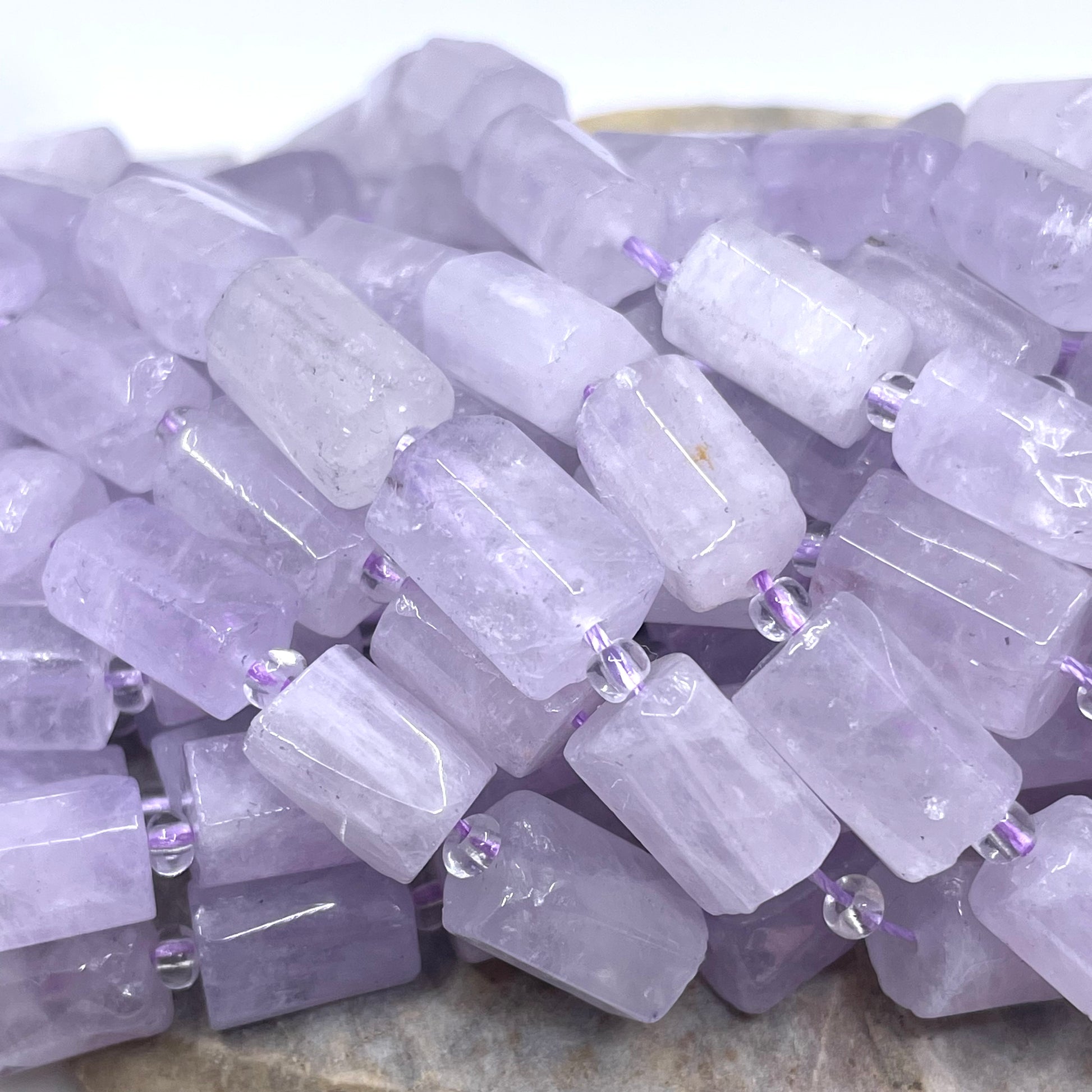 A pile of light purple amethyst rectangular gemstone beads threaded on a string, resting on a neutral stone surface.