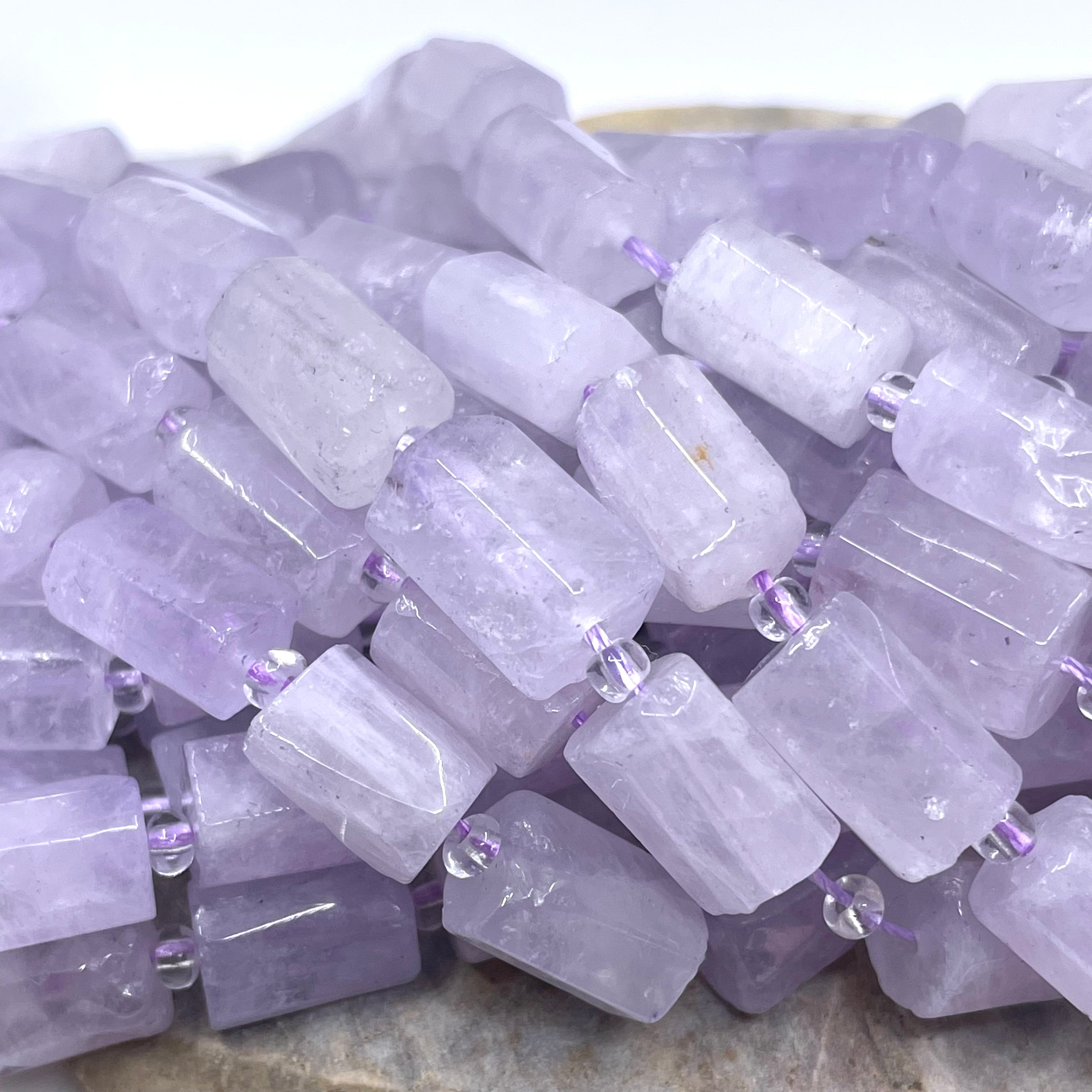 A pile of light purple amethyst rectangular gemstone beads threaded on a string, resting on a neutral stone surface.