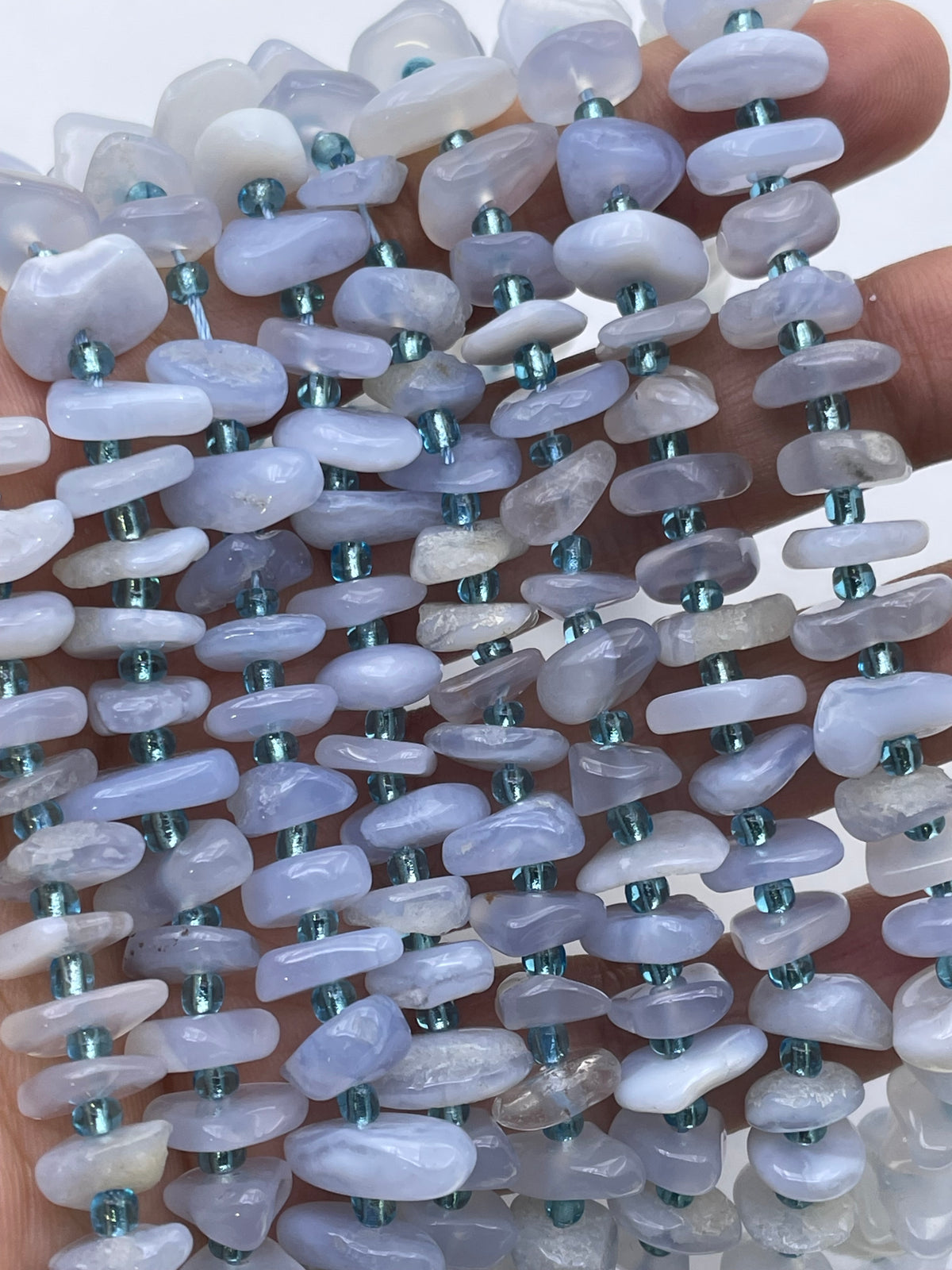 A close-up of multiple strands of irregular, polished blue lace agate stone beads in pale lavender, white, and gray, threaded with small metallic teal beads, held in a hand.
