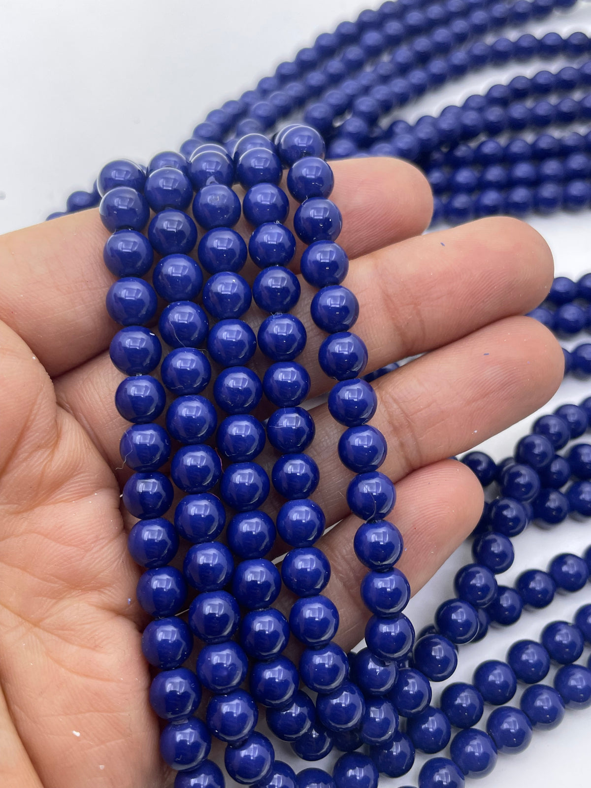 A hand holding several strands of polished, round cobalt blue shell pearl beads, with more strands draped in the background against a plain white surface.