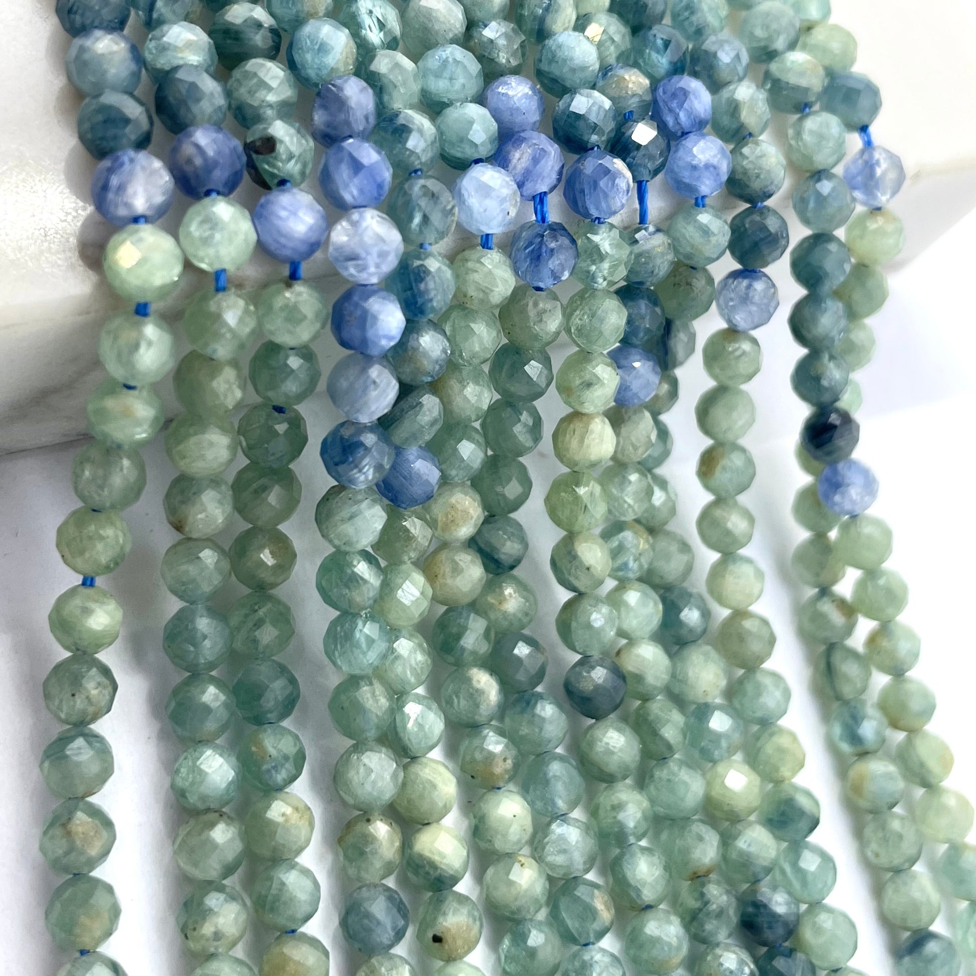 Multiple strands of faceted round rainbow green kyanite beads in shades of light green, blue, and slate gray, strung on thin blue cords and arranged vertically against a white background.