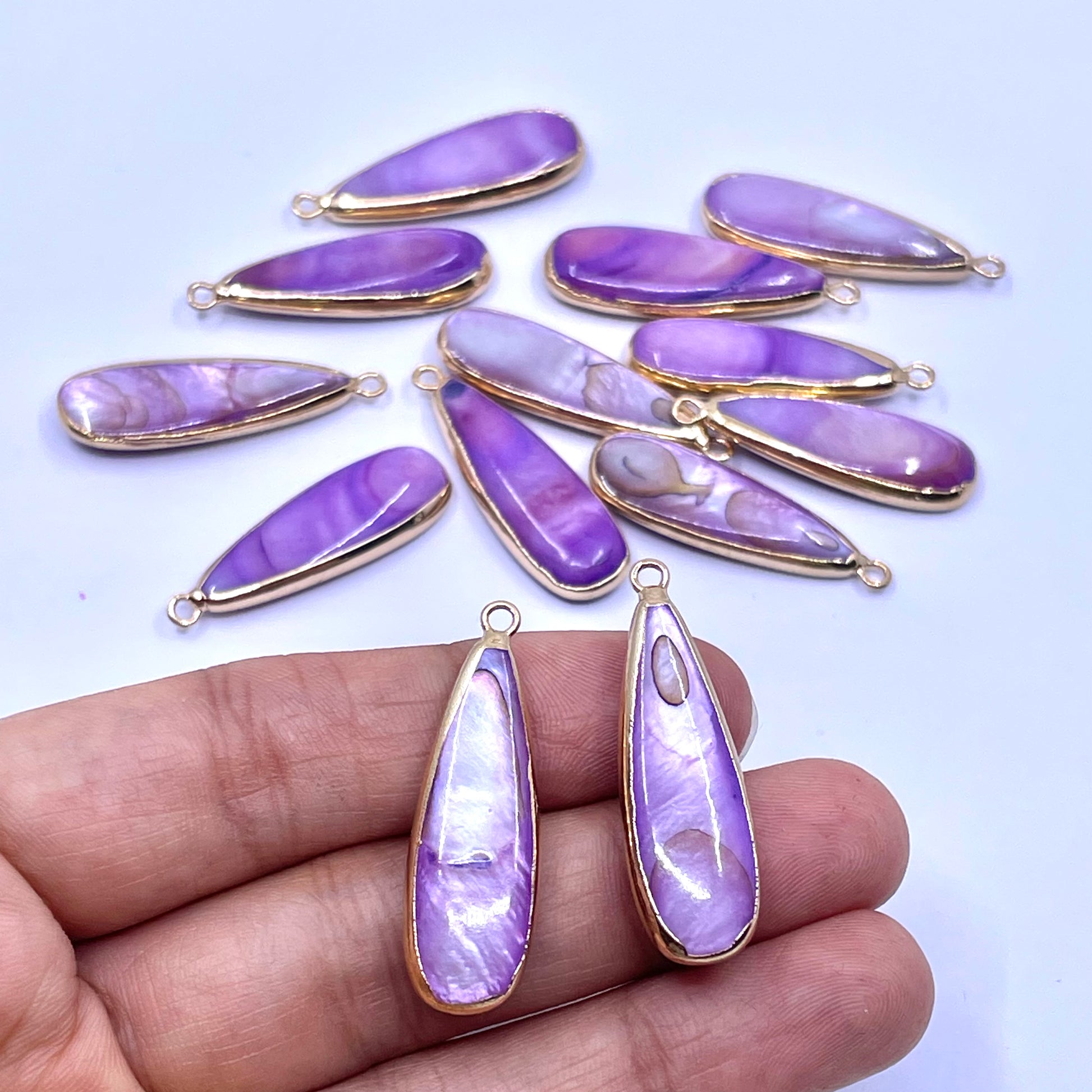 Purple teardrop-shaped pendant charms with gold-plated bezels, shown with two larger pendants held in a hand.