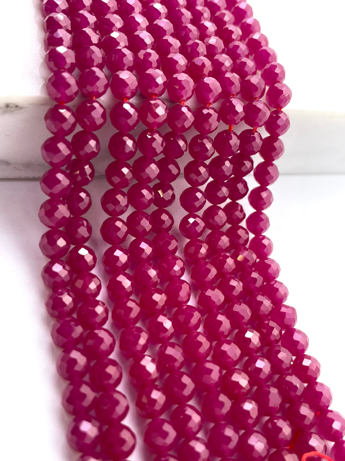 Faceted round ruby beads in a vibrant magenta-pink color, arranged in parallel strands on a white marble-like surface.