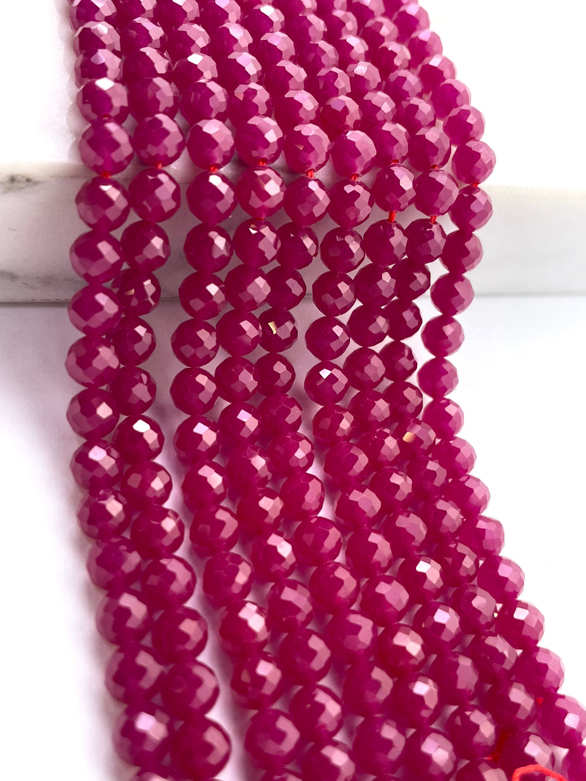 Faceted round ruby beads in a vibrant magenta-pink color, arranged in parallel strands on a white marble-like surface.