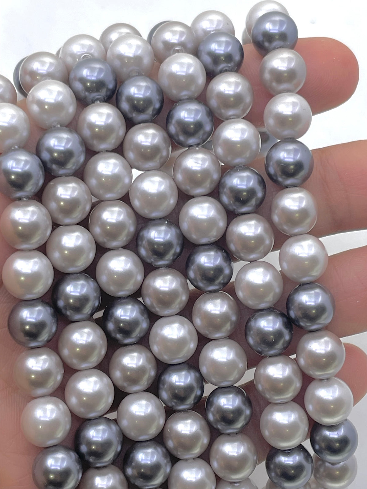A close-up of a hand holding multiple strands of round shell pearls in mixed white, gray, and silver tones.