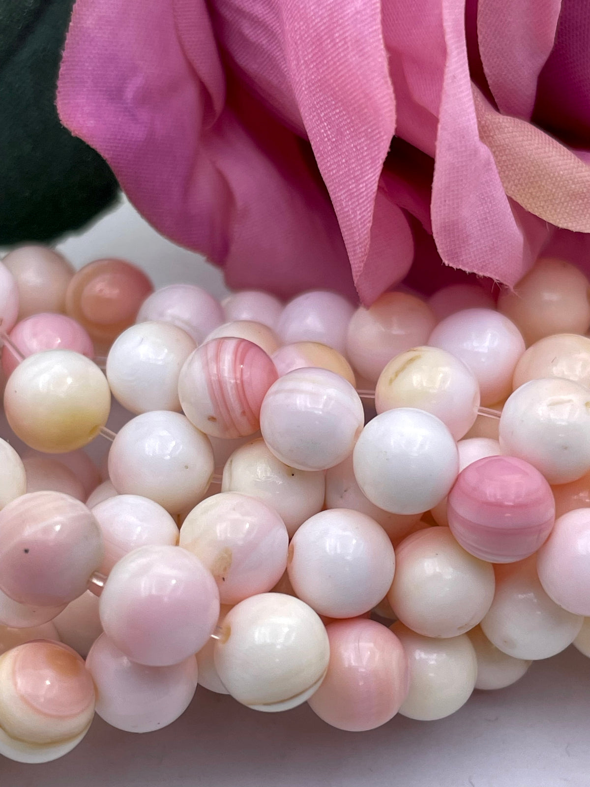 Close-up of a strand of round queen conch shell pearl beads in soft pink, white, and cream tones, with a pink fabric flower prop behind them.
