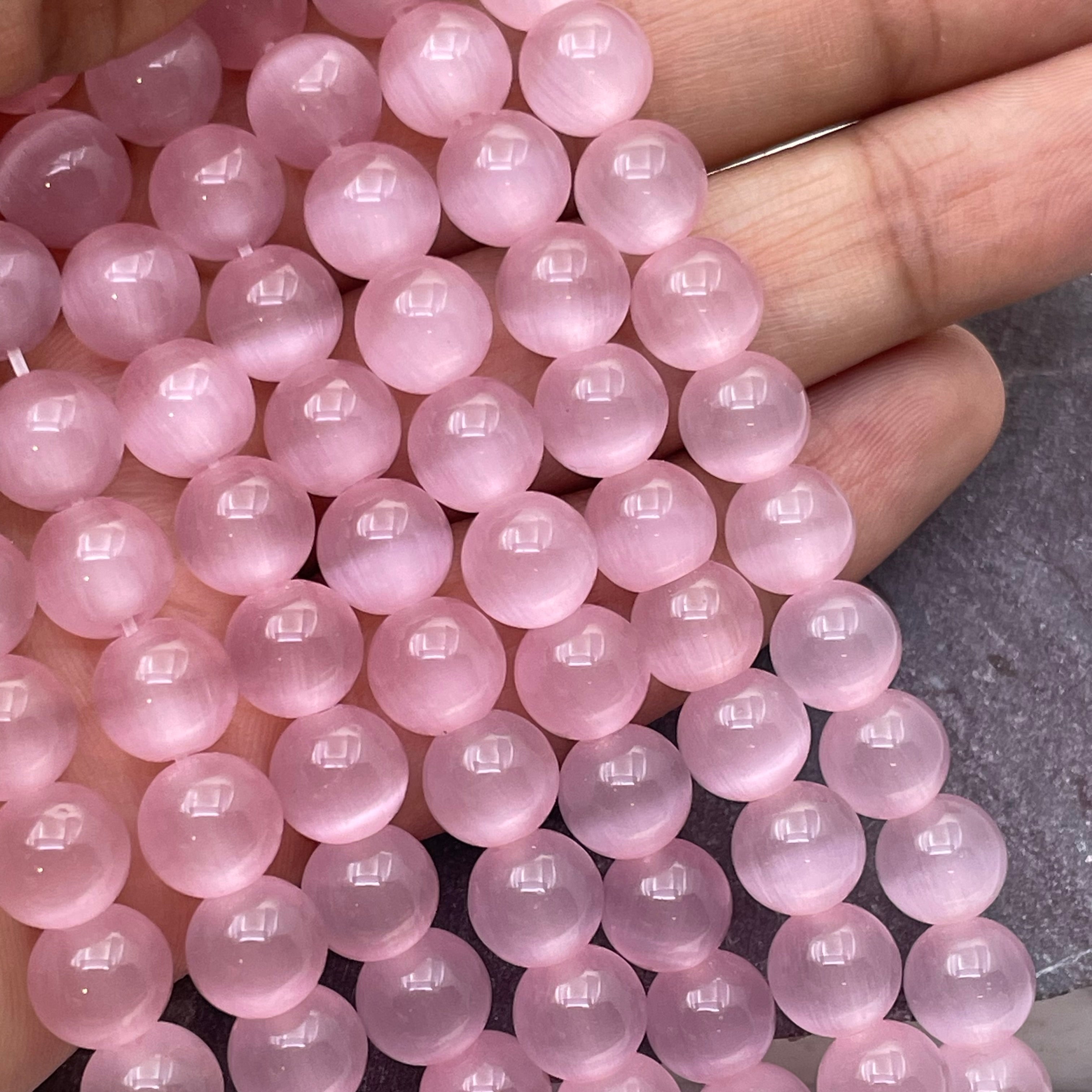 A close-up of a hand holding a strand of smooth, polished pink cats eye round beads, against a neutral gray background.