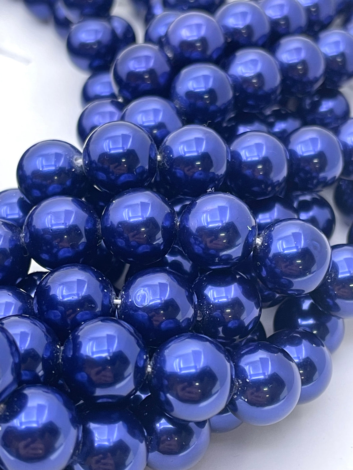 A close-up of glossy dark blue shell pearl round beads arranged in strands on a white background.
