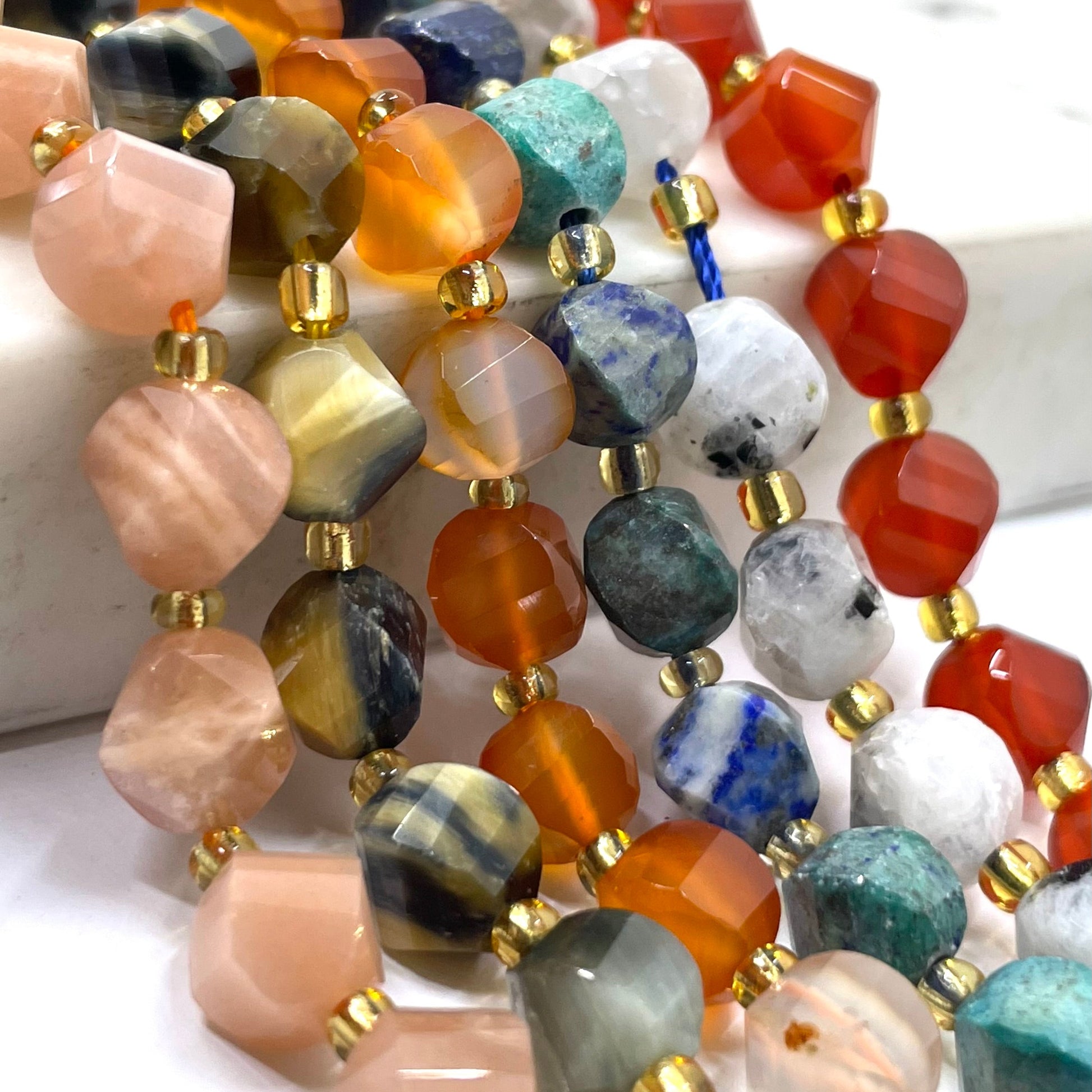 Close-up of multiple strands of faceted gemstone beads with gold spacer beads, featuring a variety of colors including orange, pink, blue, green, gray, and white.