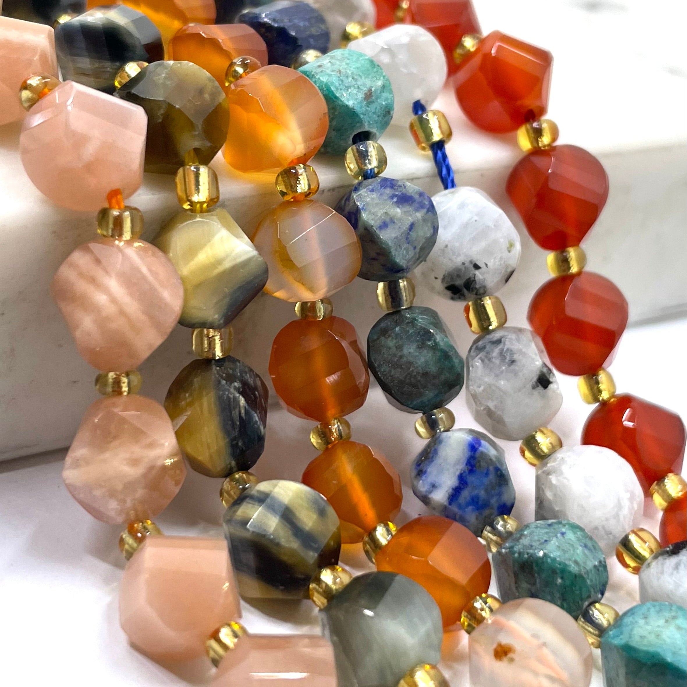 Close-up of multiple strands of faceted gemstone beads with gold spacer beads, featuring a variety of colors including orange, pink, blue, green, gray, and white.