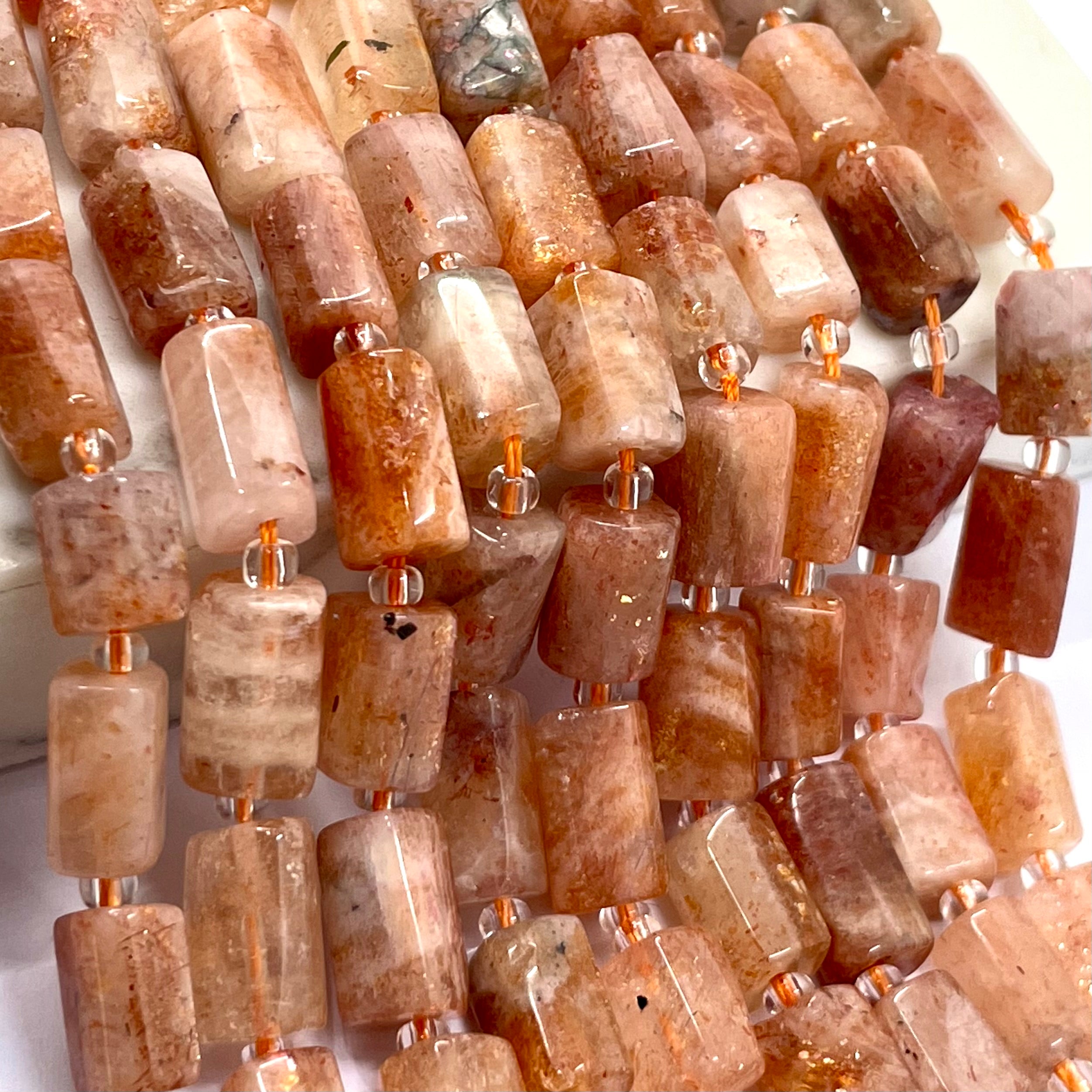 Close-up of a strand of polished rectangular sunstone gemstone beads in warm peach, orange, brown, cream, and translucent tones, separated by small clear spacer beads with metallic connectors.