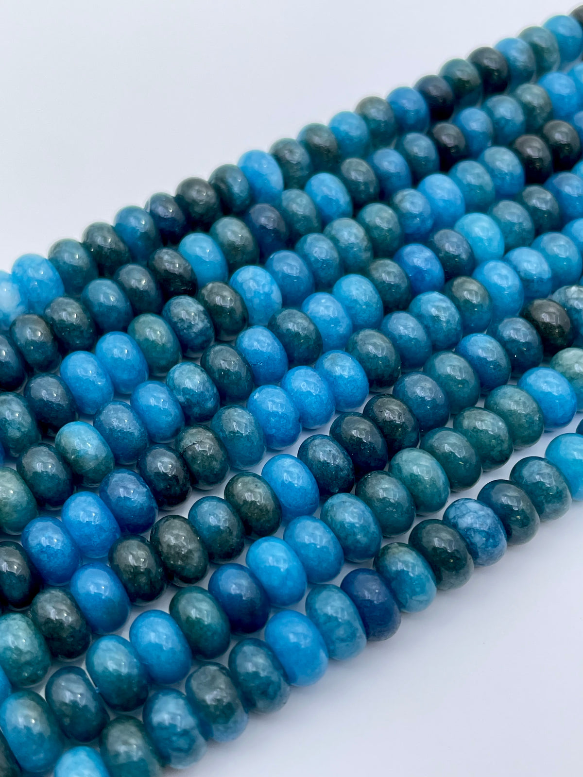 Multiple strands of smooth oval jade apatite beads in shades of blue and green, arranged diagonally on a plain white background.