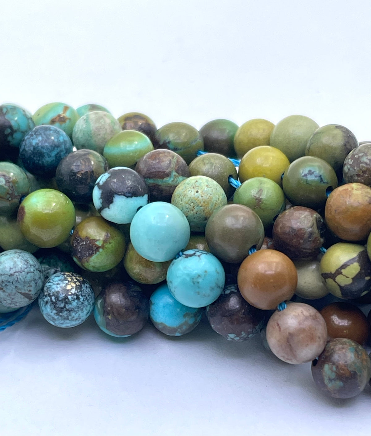 A close-up of multiple strands of round turquoise stone beads in various earthy colors including green, brown, blue, and beige, arranged on a plain white background.