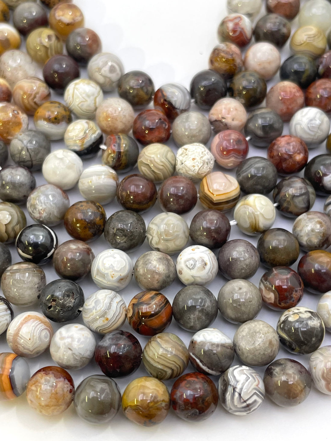A close-up of many polished round stone mexican crazy agate beads in assorted natural patterns and colors, including gray, brown, cream, black, and reddish tones, arranged on a white background.